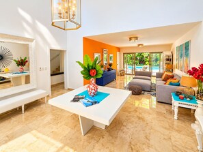 A modern living room with a white coffee table, a blue sofa, and a grey armchair. There is a large mirror on the wall, a chandelier, and a view of a pool through the glass doors. - Villa Caribe Tortuga B40 (Punta Cana)
