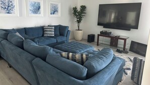Ein modernes Wohnzimmer mit einem großen, blauen Ecksofa, einem Flachbildfernseher, einer Topfpflanze und gerahmten Kunstwerken an der Wand.