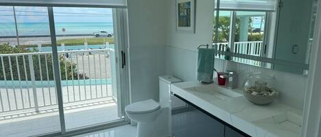 A modern bathroom with a toilet, sink, and large mirror. There is a view of the ocean through the sliding glass door.