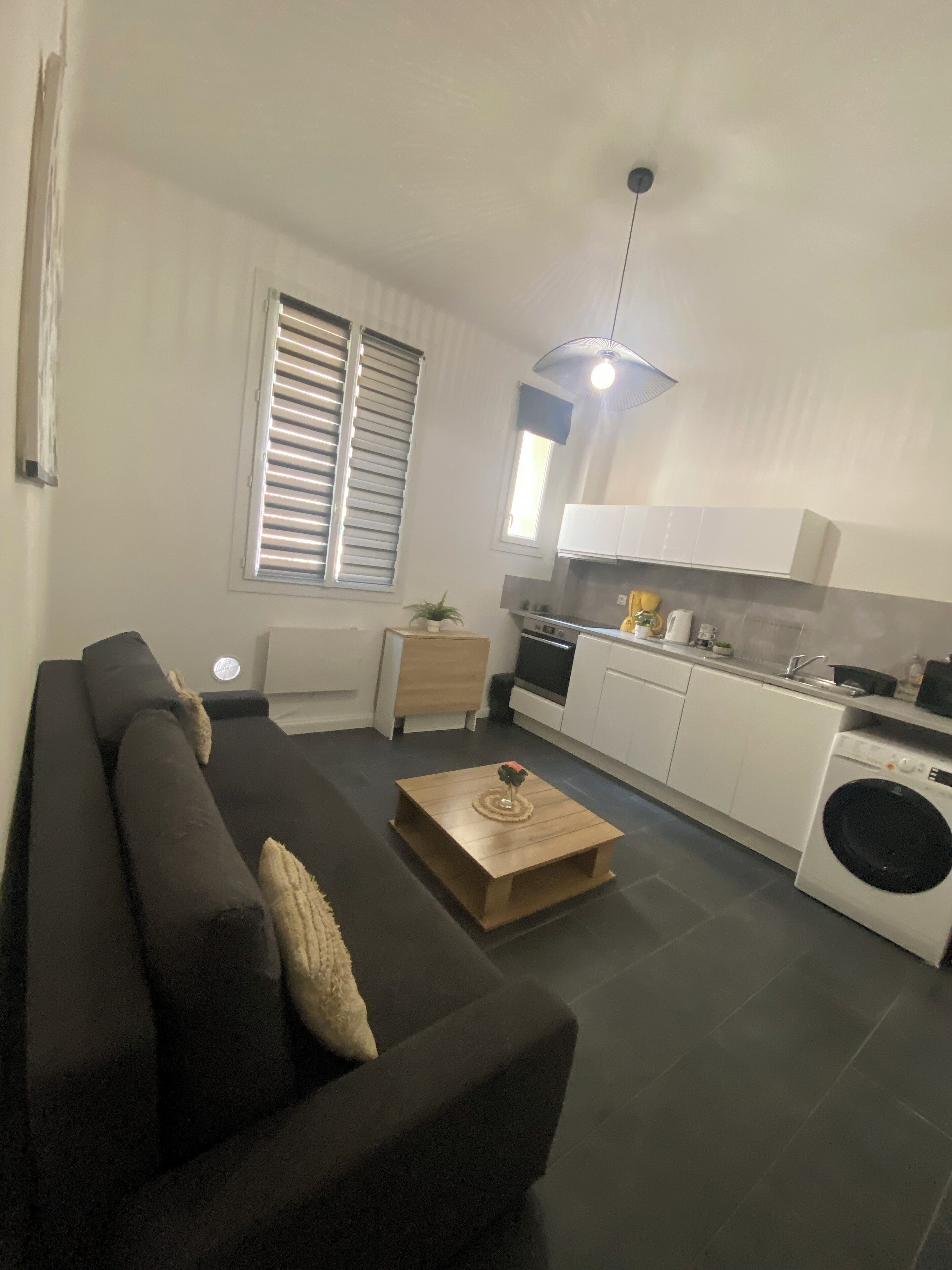 Un salon moderne avec un canapé noir, une table basse en bois et un coin cuisine doté de meubles blancs et d’un lave-linge.