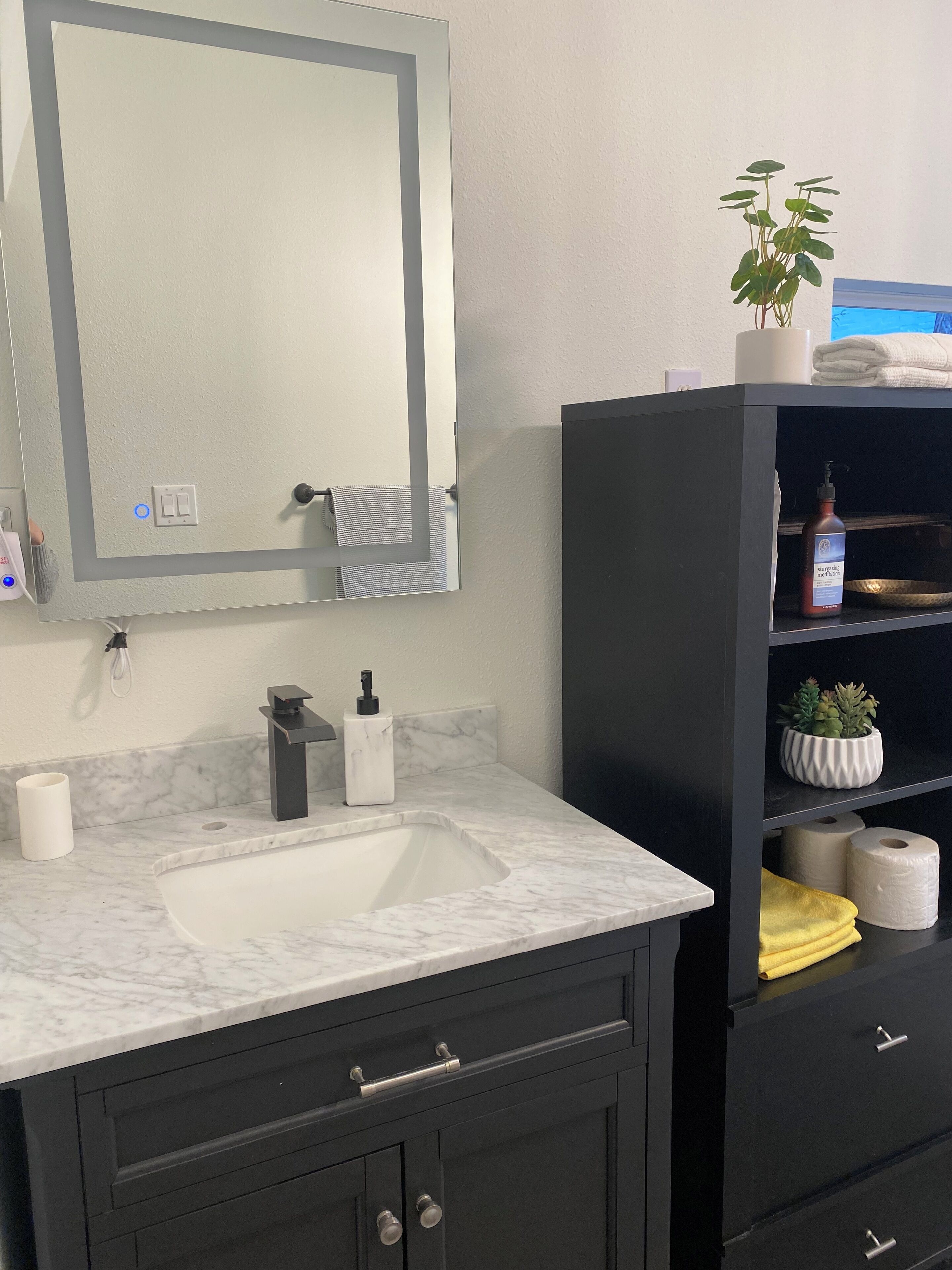 A bathroom with a marble sink, a black vanity with drawers, a mirror, and a shelf with toiletries.
