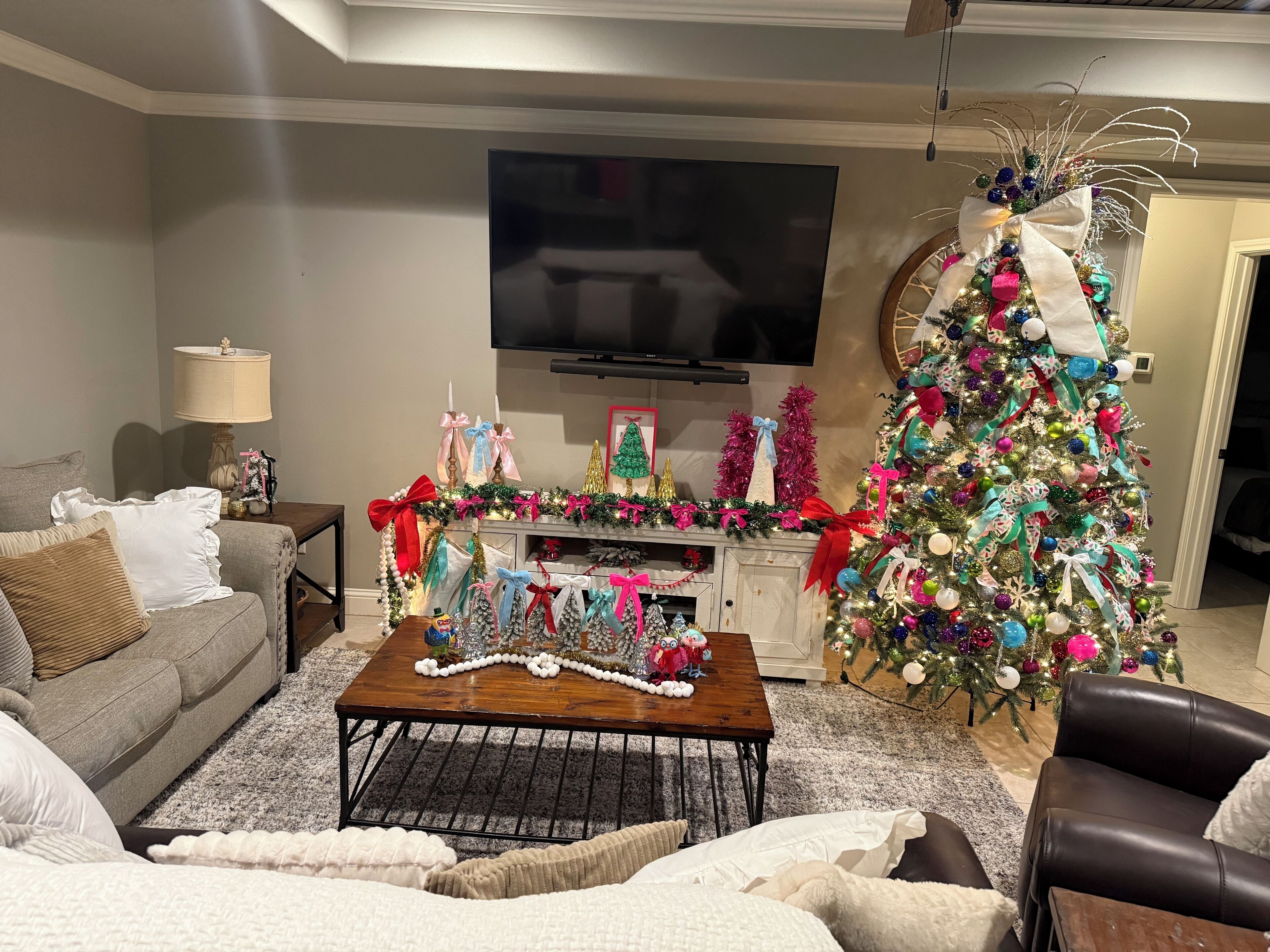 A living room with a large Christmas tree, a flat-screen TV, and a fireplace mantel decorated with festive ornaments.