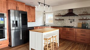 A modern kitchen with wooden cabinets, a stainless steel refrigerator, a wooden island with stools, and a herringbone-patterned backsplash. - HAVEN COTTAGE - Close to Mpls, Far From Your Routine-FREE SUPS on Lake Fremont! (Zimmerman)