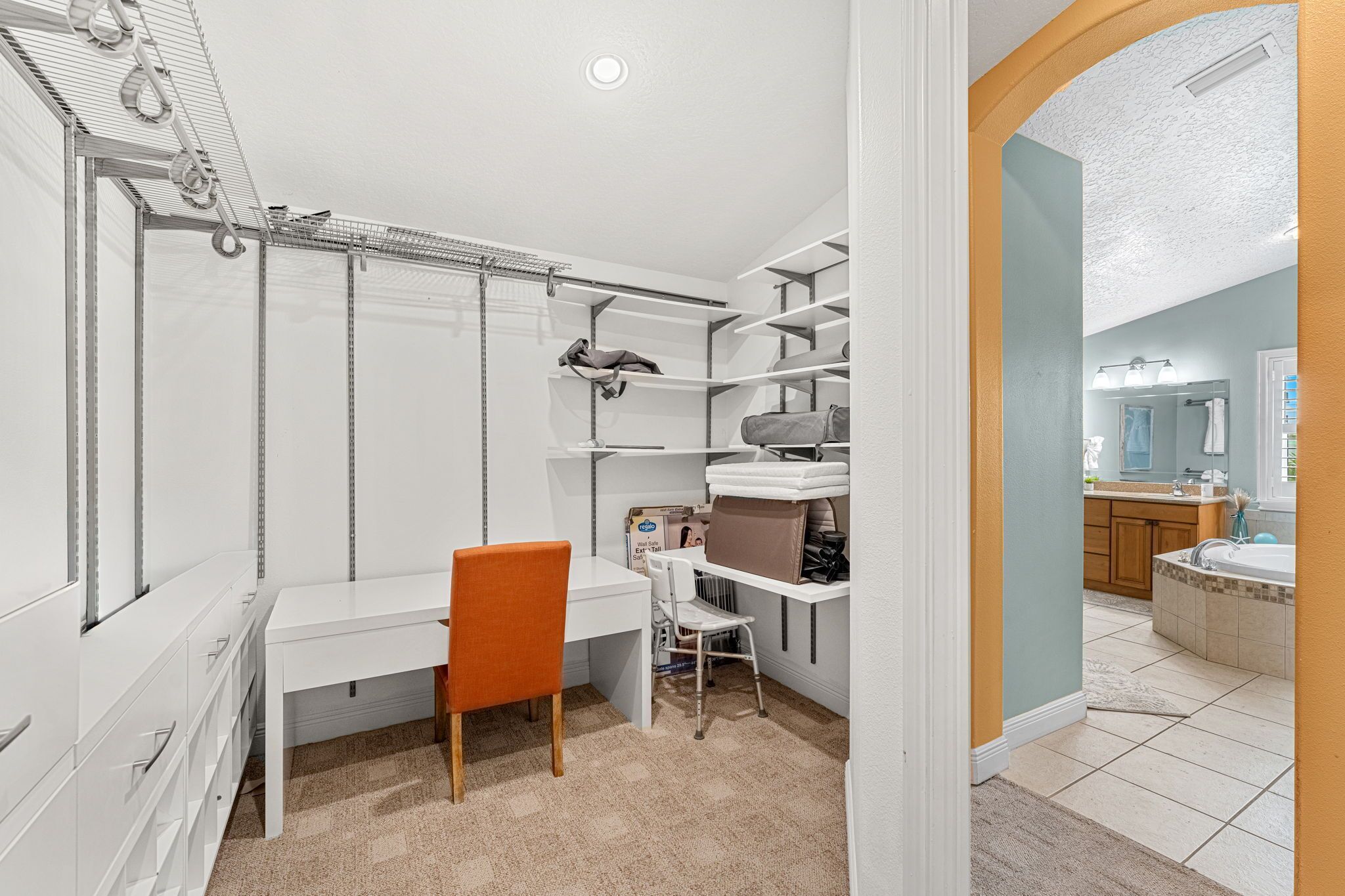 Une salle de bain compacte comprenant une douche, des toilettes et un lavabo. On y trouve un petit bureau avec une chaise et une étagère contenant des articles de toilette. La pièce est dotée d'un carrelage au sol et d'une fenêtre avec des stores.