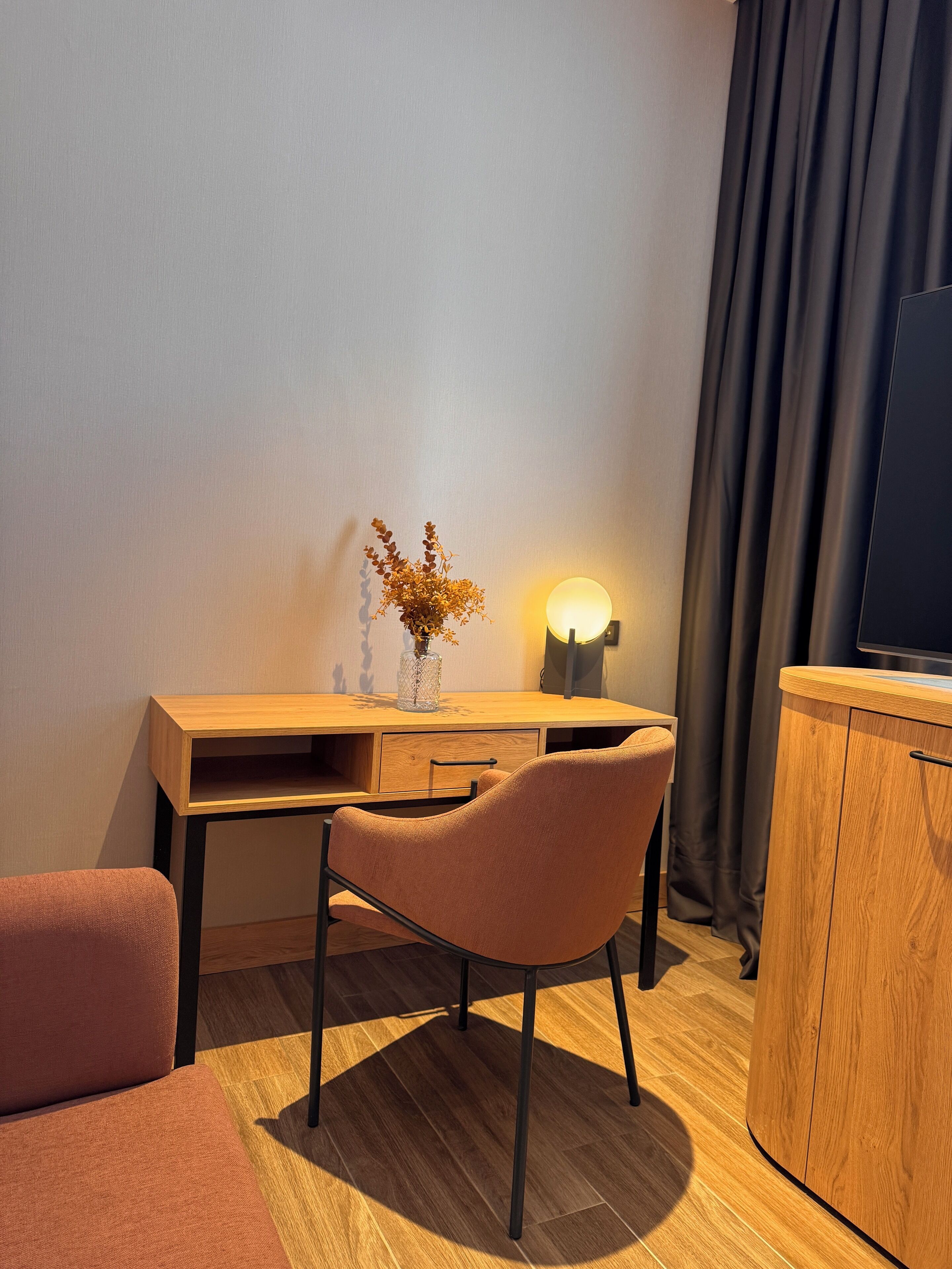 Une chambre moderne avec un bureau en bois, une chaise rembourrée, un petit vase contenant des fleurs séchées, une lampe fixée au mur, un téléviseur et un rideau foncé.