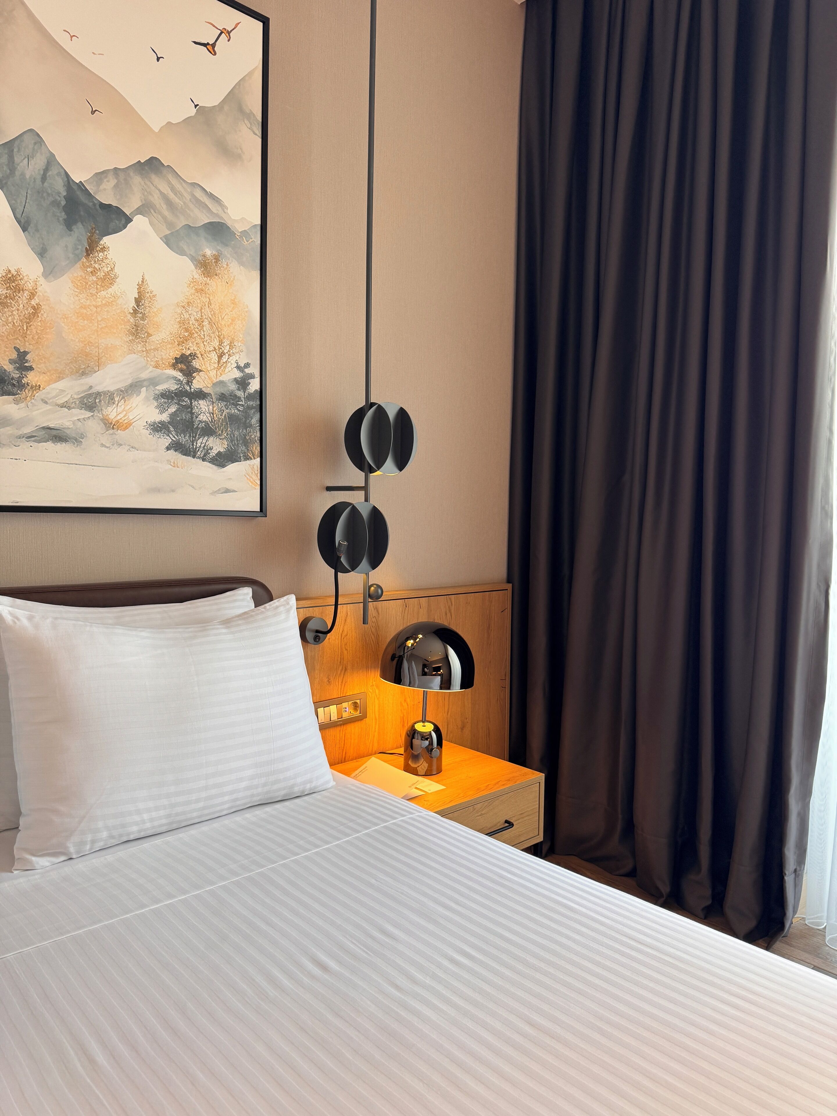 A neatly made bed with white linens, a wooden nightstand with a lamp, and a painting of mountains and trees on the wall.