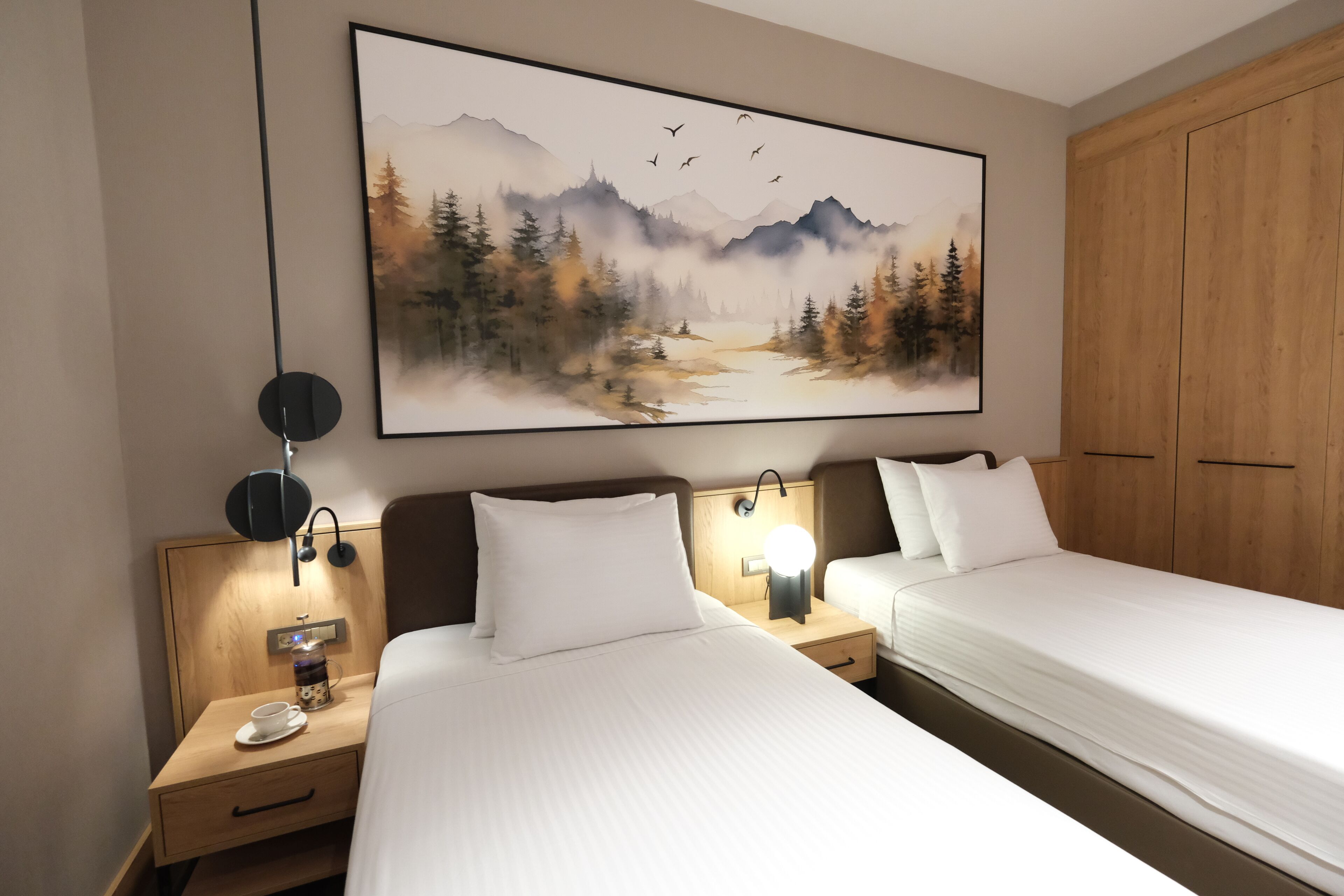 Hotellihuone, jossa on kaksi sänkyä, suuri maalaus, jossa on kuvattuna metsä ja vuoristo, puinen vaatekaappi sekä yöpöytä, jossa on lamppu ja kuppi.