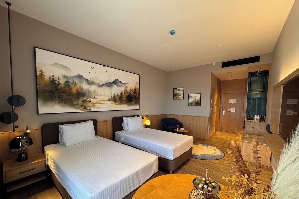 A hotel room with two beds, a dining table with a fruit platter, a painting of a forest, and a view of the hallway.