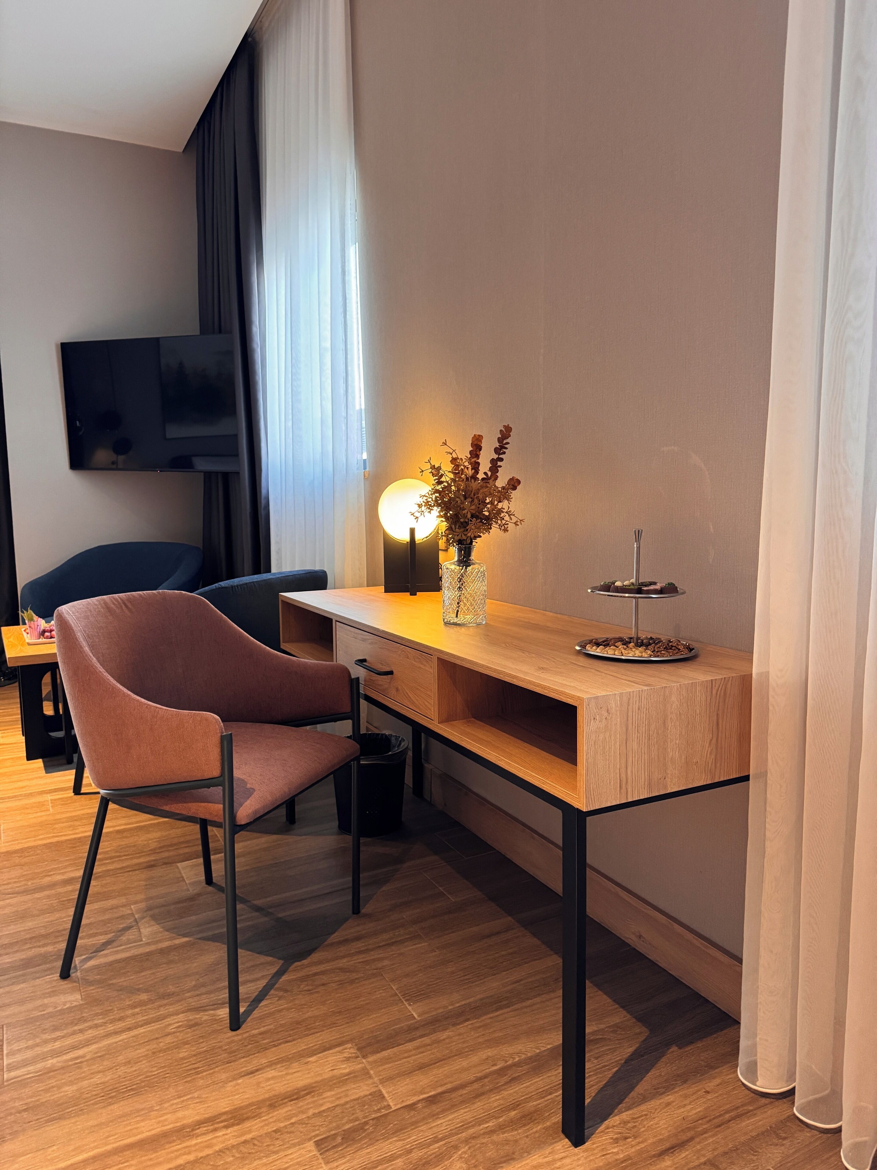 A modern hotel room with a wooden desk, a comfortable armchair, a television mounted on the wall, and a vase with dried flowers on the desk.
