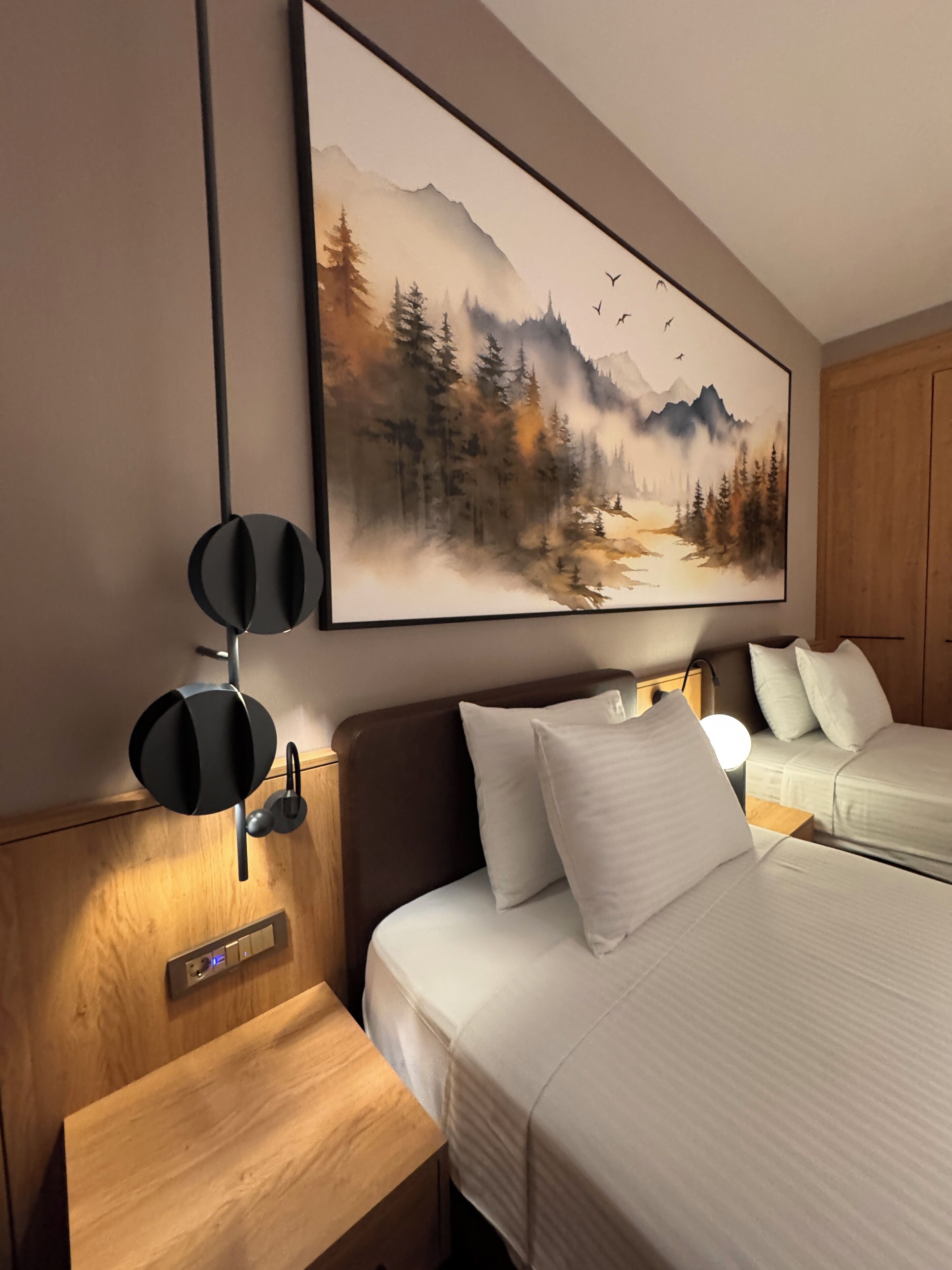A hotel room with a large painting of a forest and mountains, a wooden headboard, two beds, and a bedside table with a lamp.