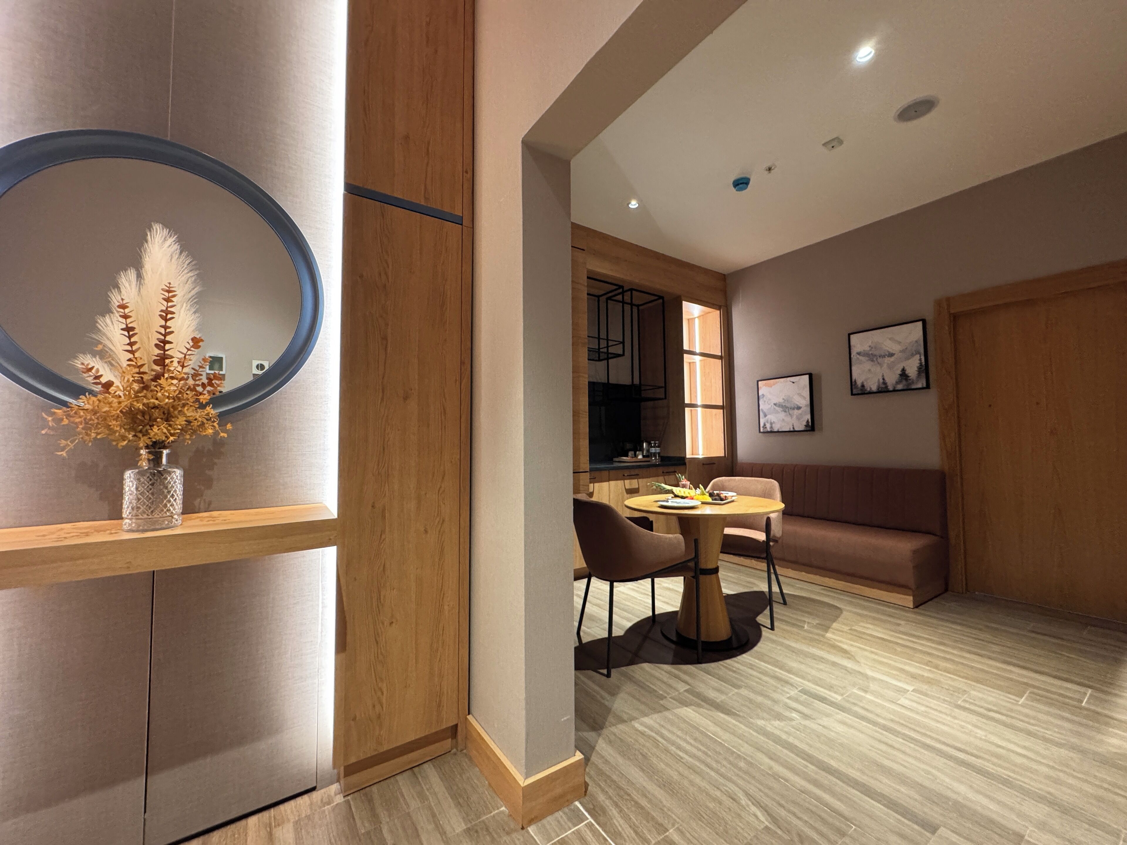 A modern interior with a wooden dining table, a round mirror, and a vase with dried flowers.