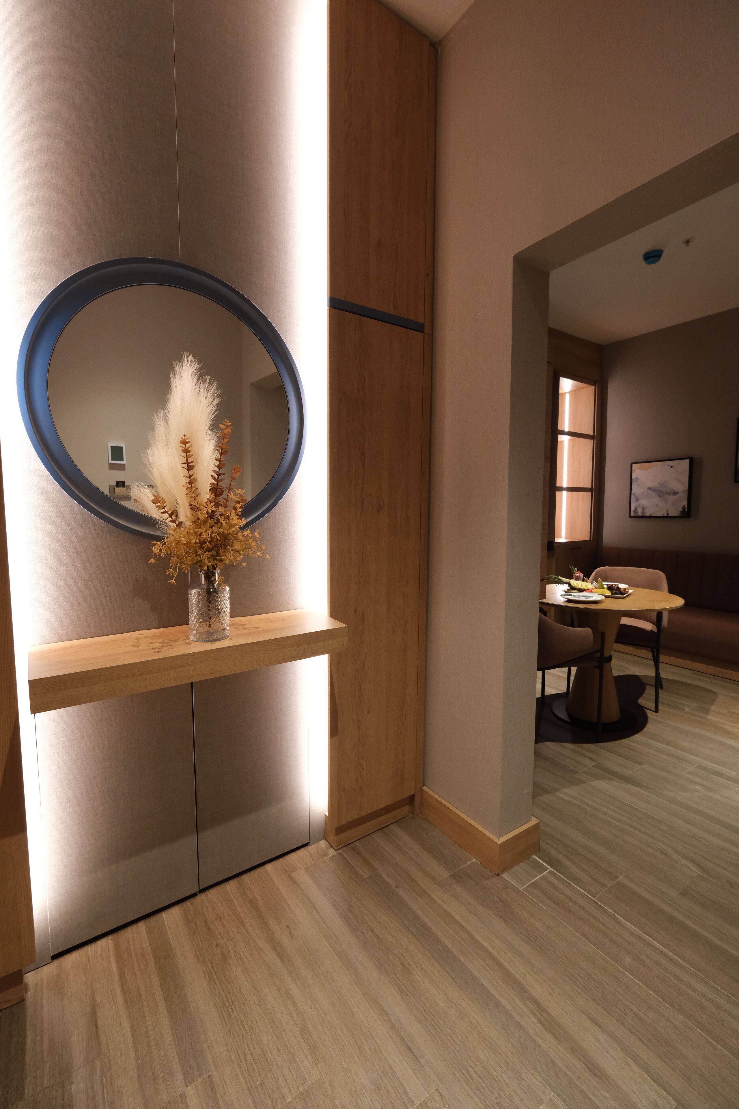 A modern hotel room with a wooden floor, a round mirror with a blue frame, a vase with dried flowers, and a dining area with a table and chairs.