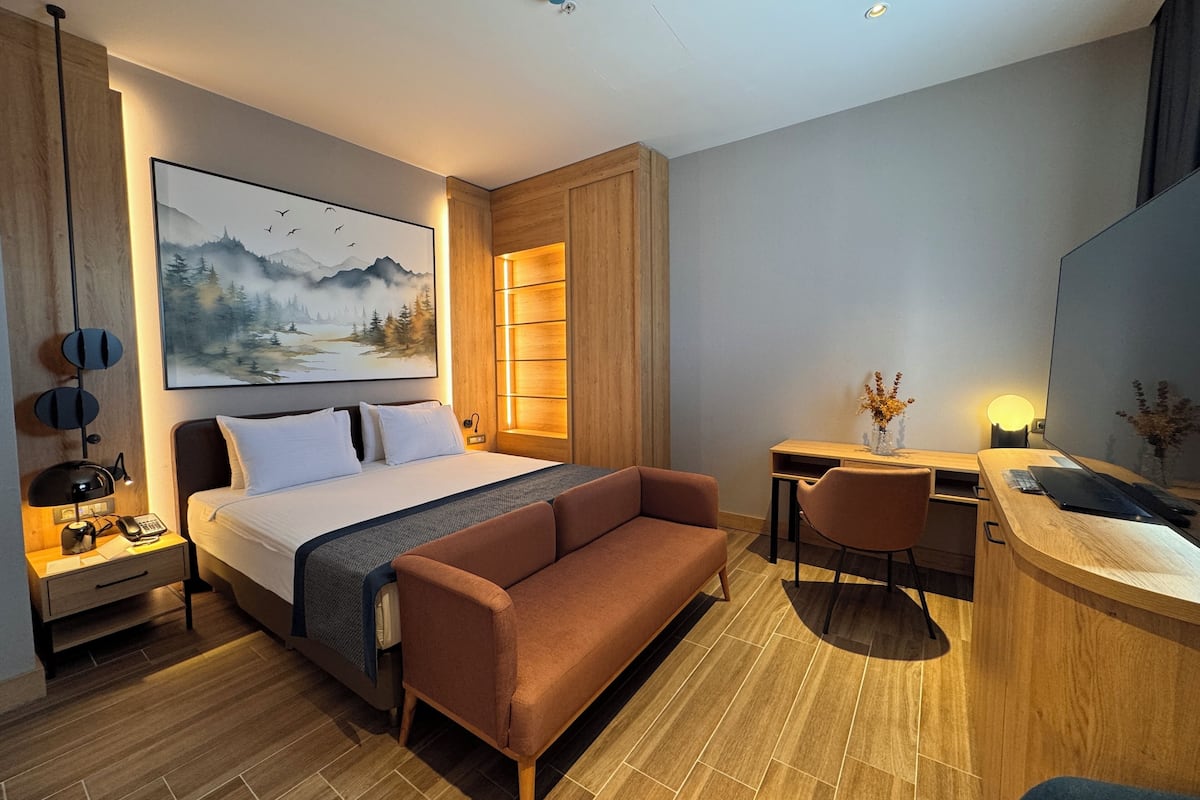 A modern hotel room with a large bed, a sofa, a desk with a computer, and a bathroom area.