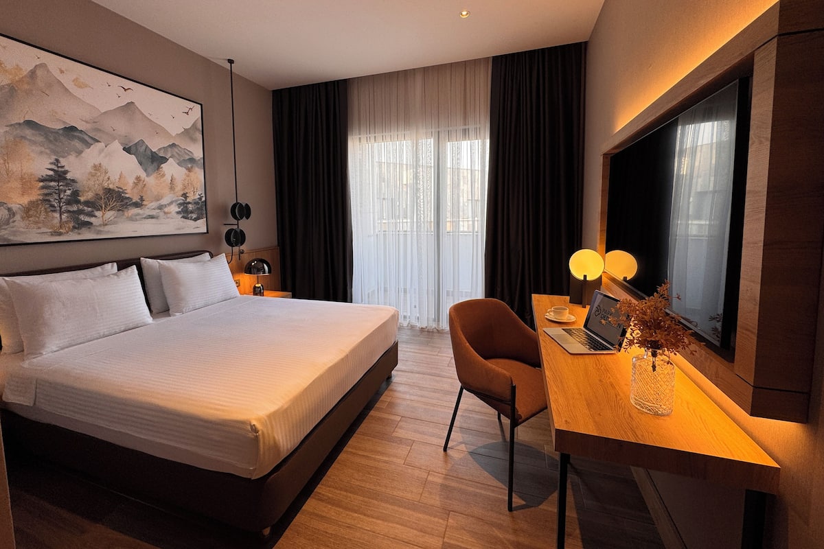 A hotel room with a large bed, a desk with a laptop, a chair, a TV, and a painting on the wall.