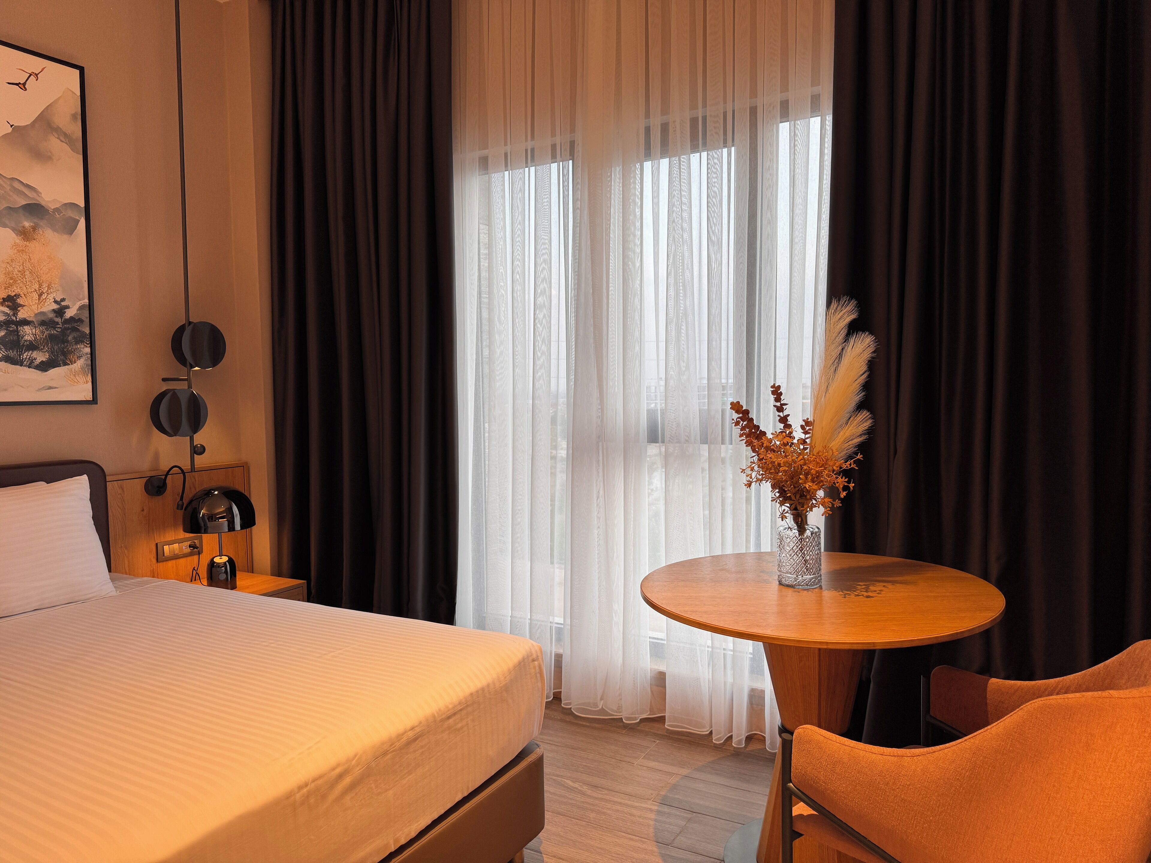 Une chambre d’hôtel avec un lit, une table ronde ornée d’un vase de fleurs séchées, une chaise, une table de chevet avec une lampe et un tableau accroché au mur.