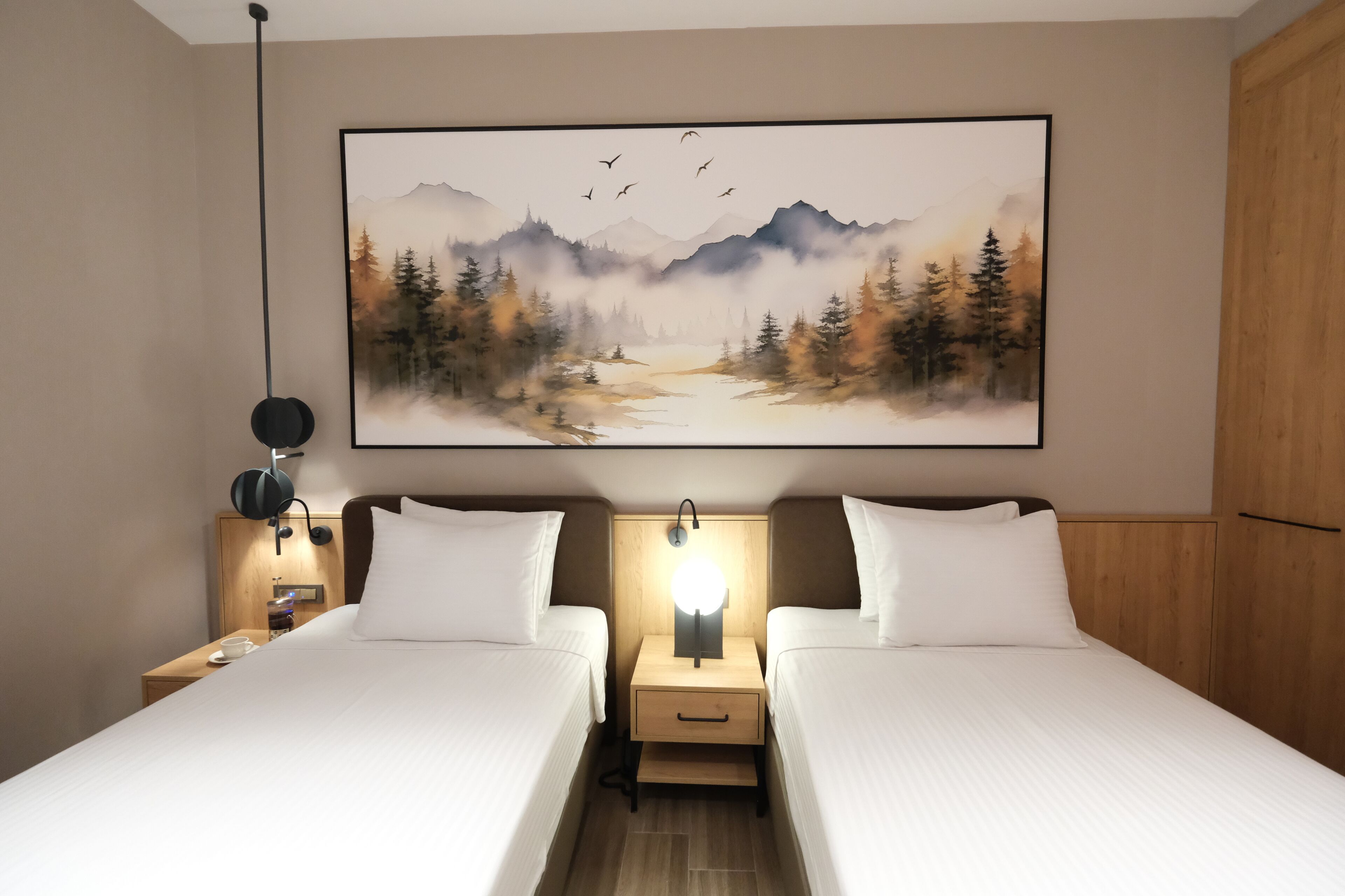 A hotel room with two beds, a nightstand with a lamp, a painting of a forest scene, and a wooden wardrobe.