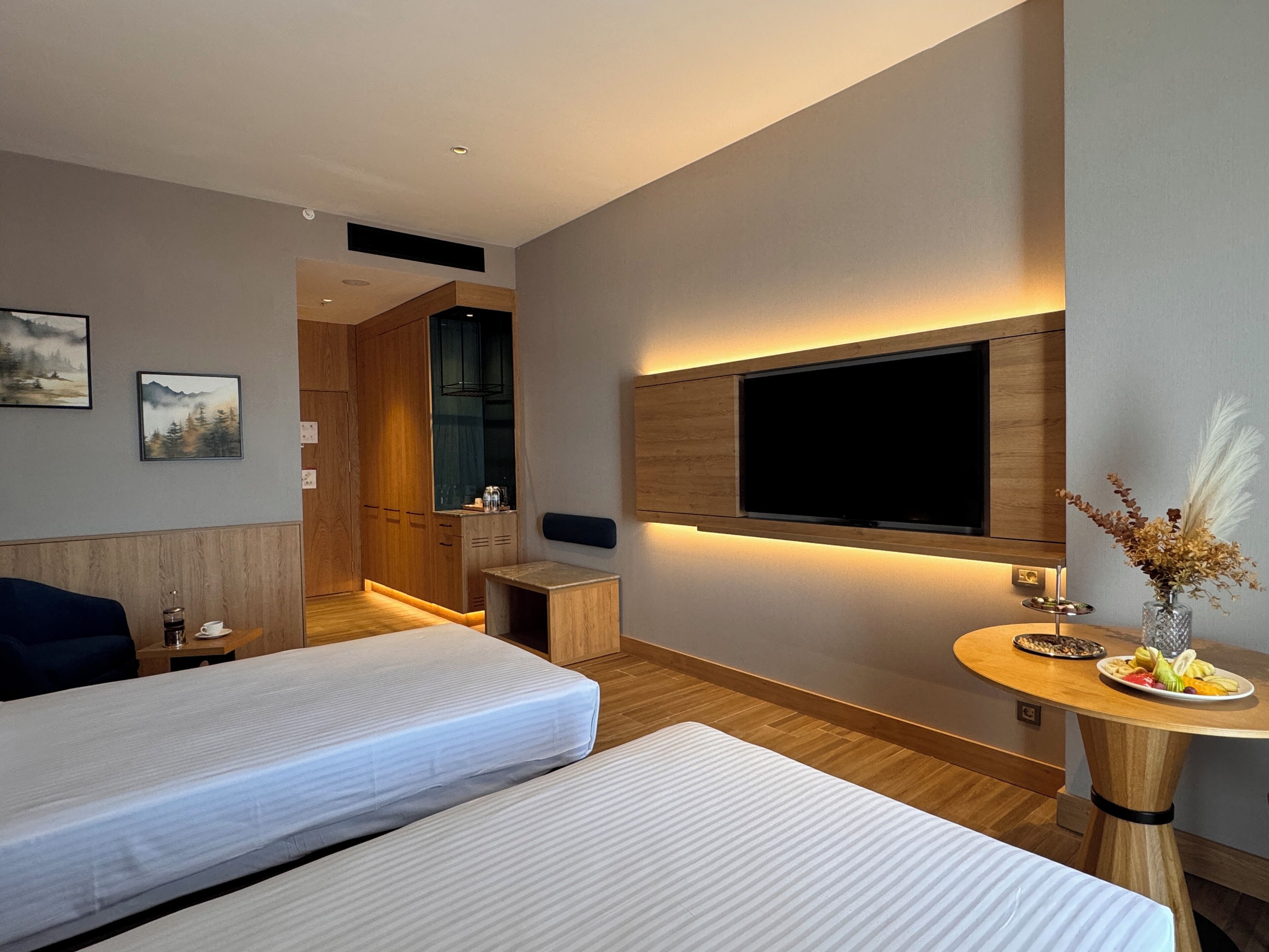 A modern hotel room with two beds, a TV mounted on the wall, a small table with a fruit plate, and a wooden cabinet.