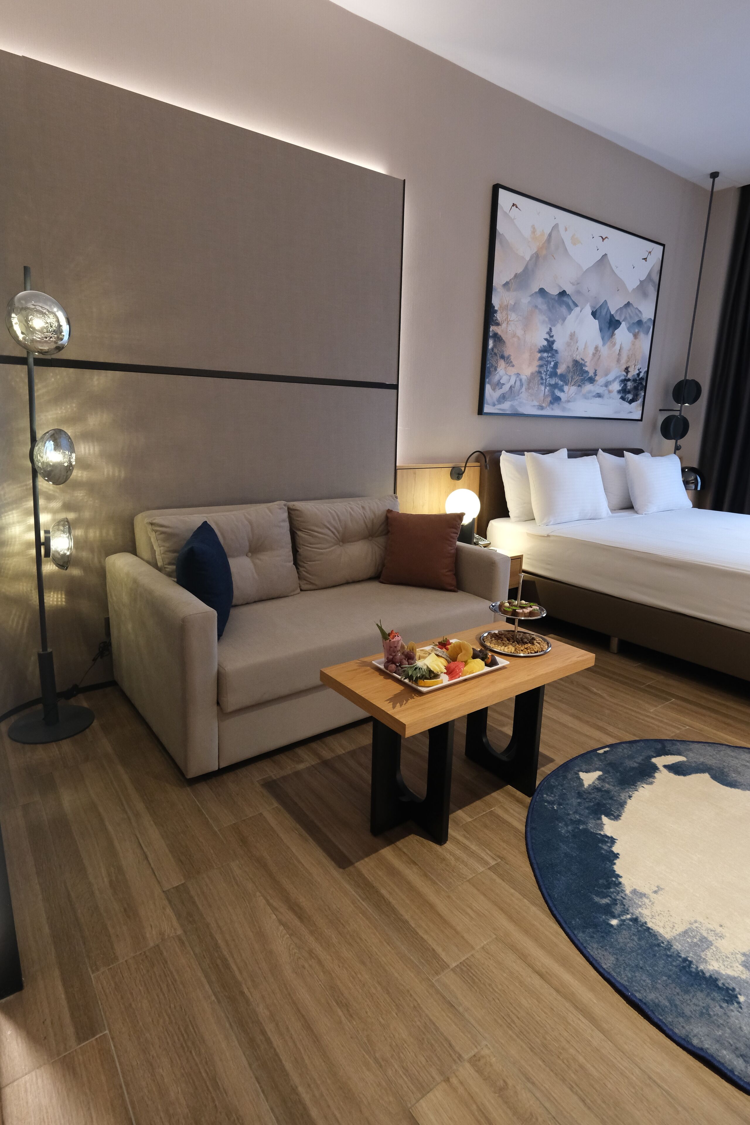 Una habitación de hotel moderna con sofá, una mesa de comedor con fruta, cama y un cuadro en la pared.