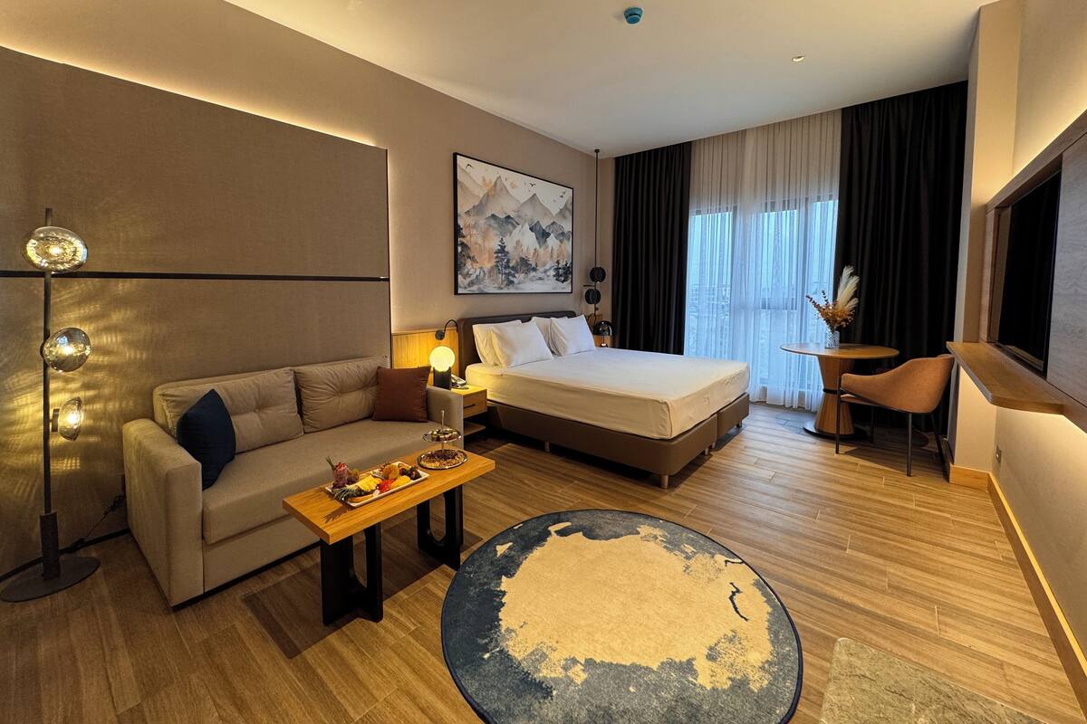 A modern hotel room with a bed, sofa, desk, and a large window with curtains.