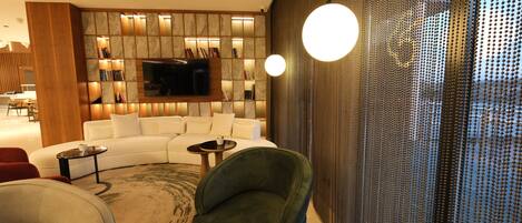 Una sala moderna con un sofá seccional, un sillón verde, una mesa de centro redonda y un televisor montado en una pared con textura de piedra.