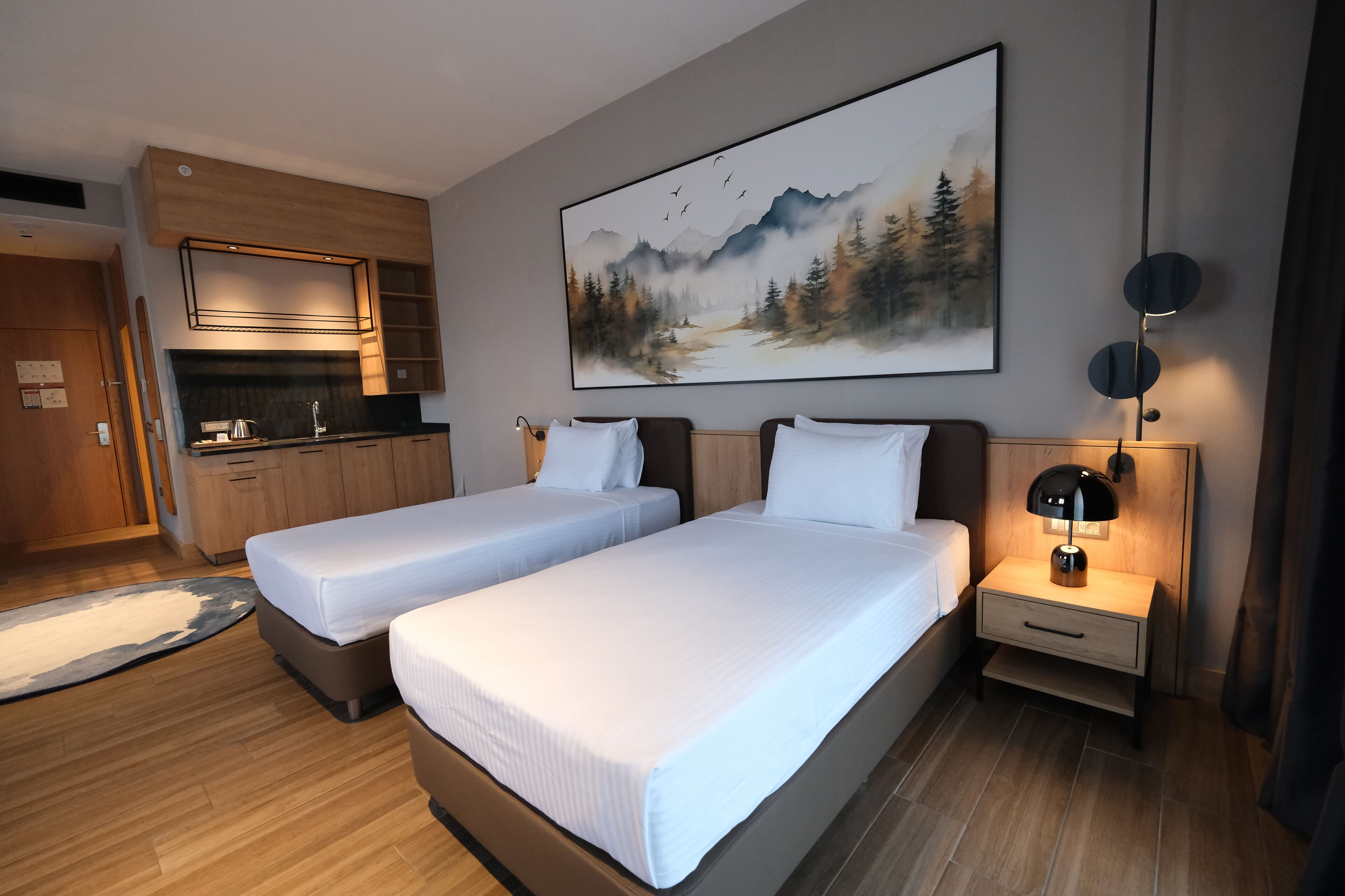 Quarto de hotel com duas camas, uma kitchenette pequena, um quadro com cena de floresta e uma mesa de cabeceira com abajur.