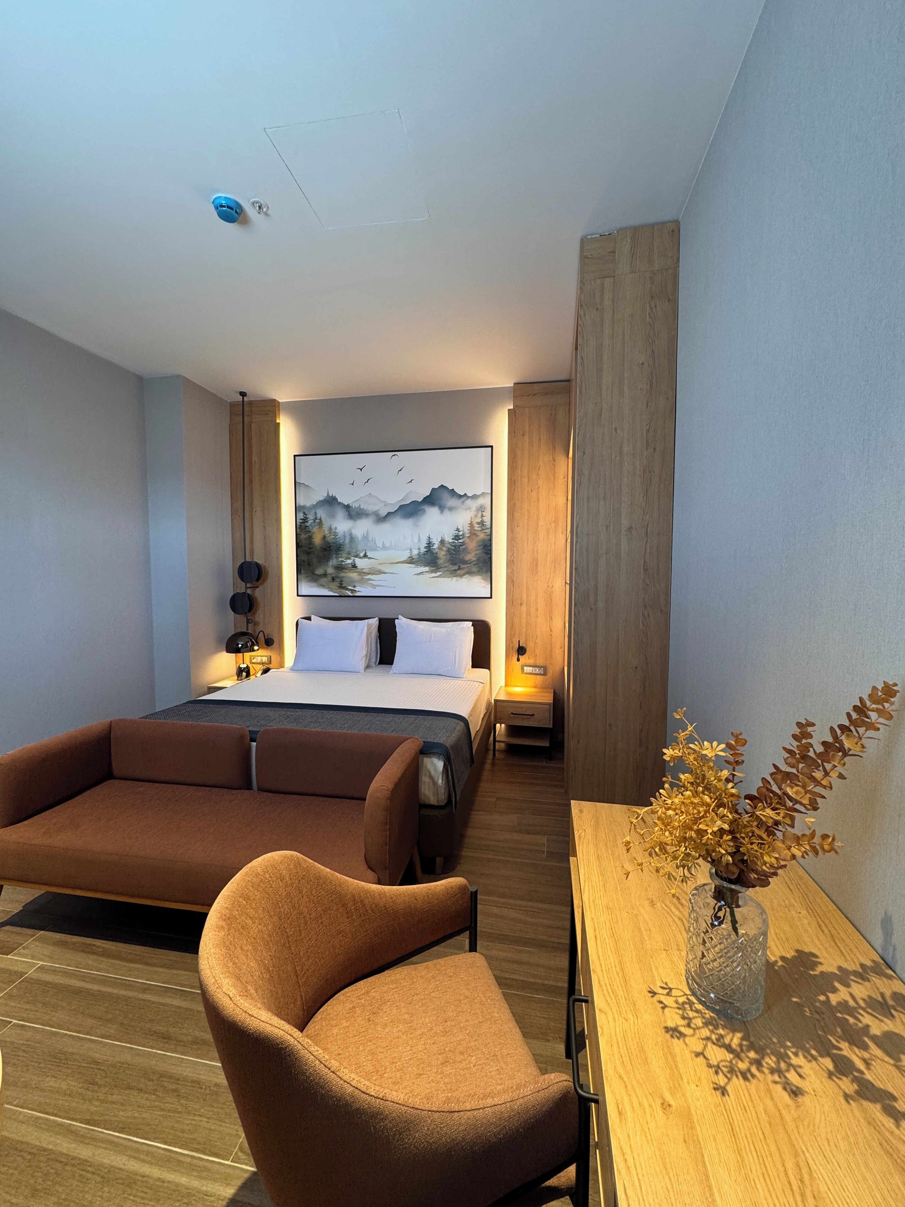 A modern hotel room with a large bed, a sofa, a small table with a vase, and a painting of mountains on the wall.
