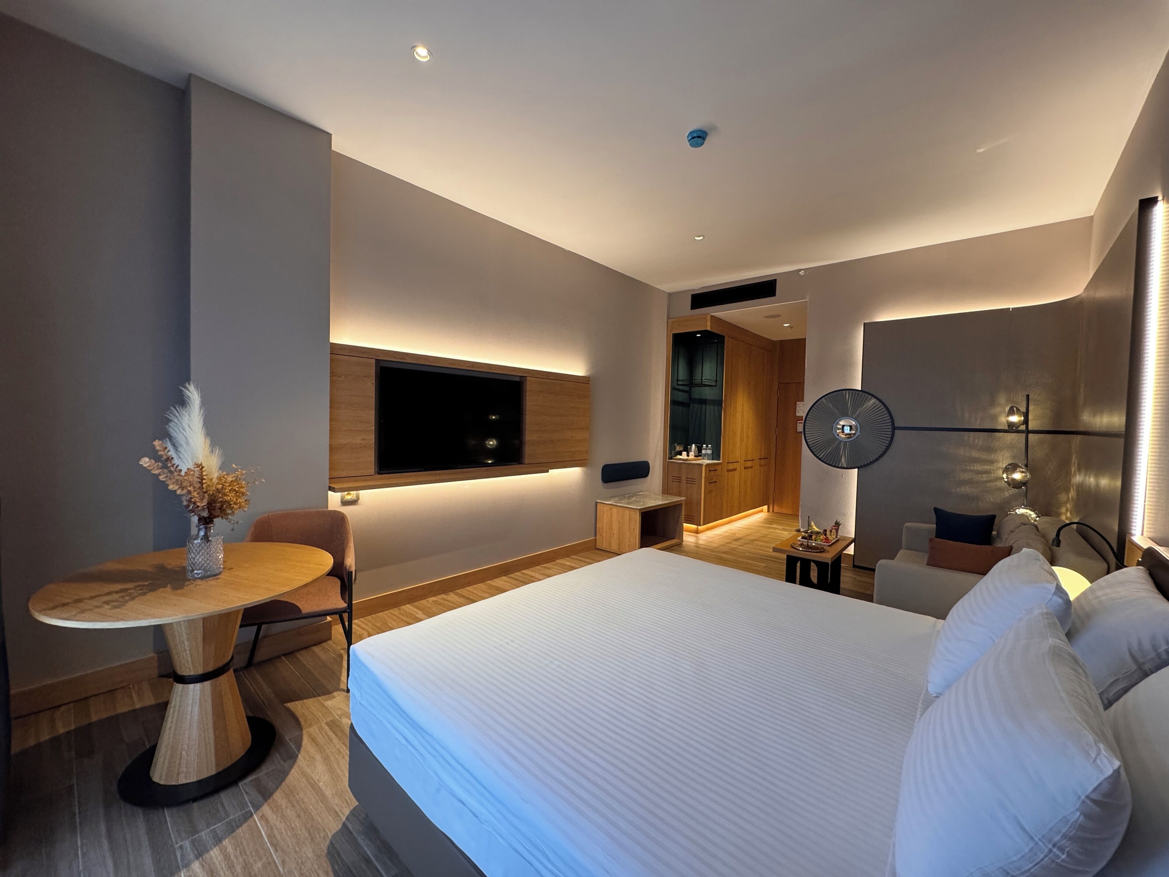 Una habitación de hotel moderna con una cama grande, un televisor montado en la pared, una pequeña mesa de comedor y un rincón de descanso con un sofá y sillones.