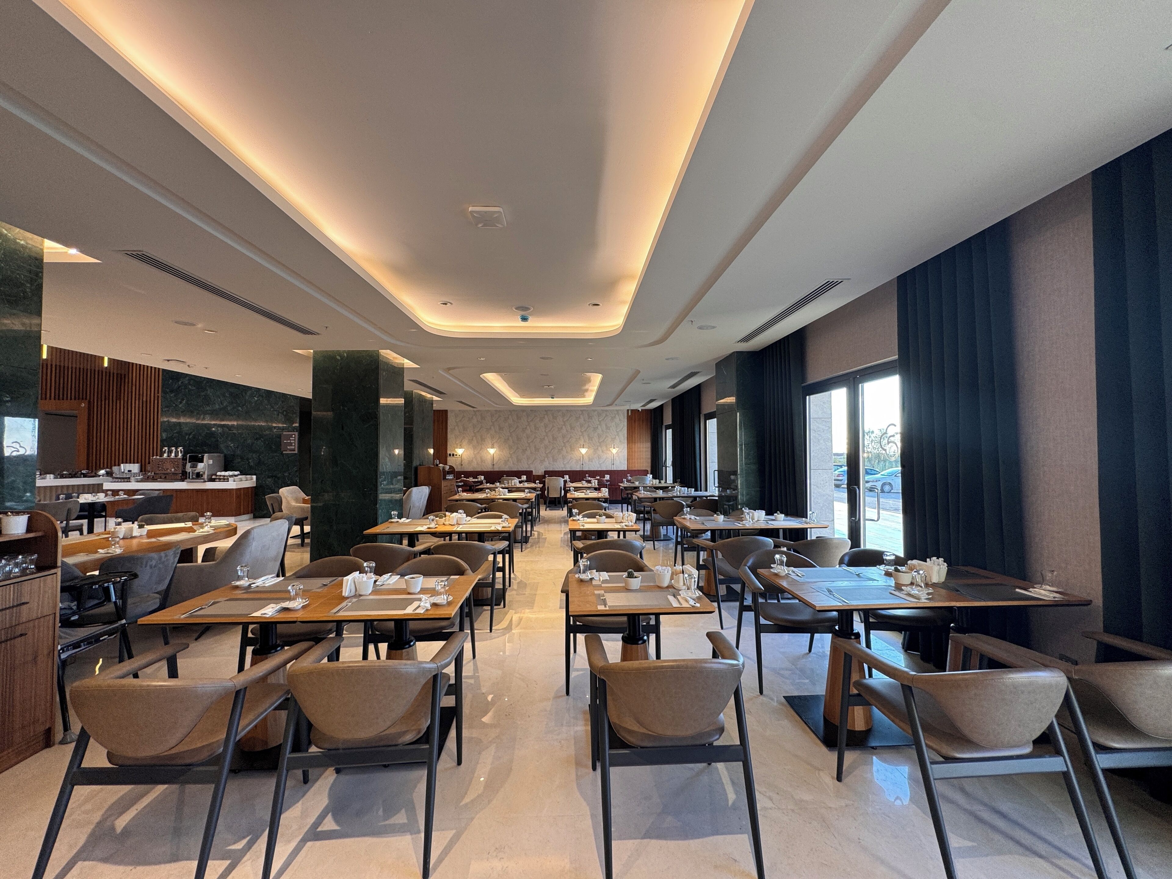 A modern dining area with multiple tables and chairs, a kitchen area, and large windows providing natural light.