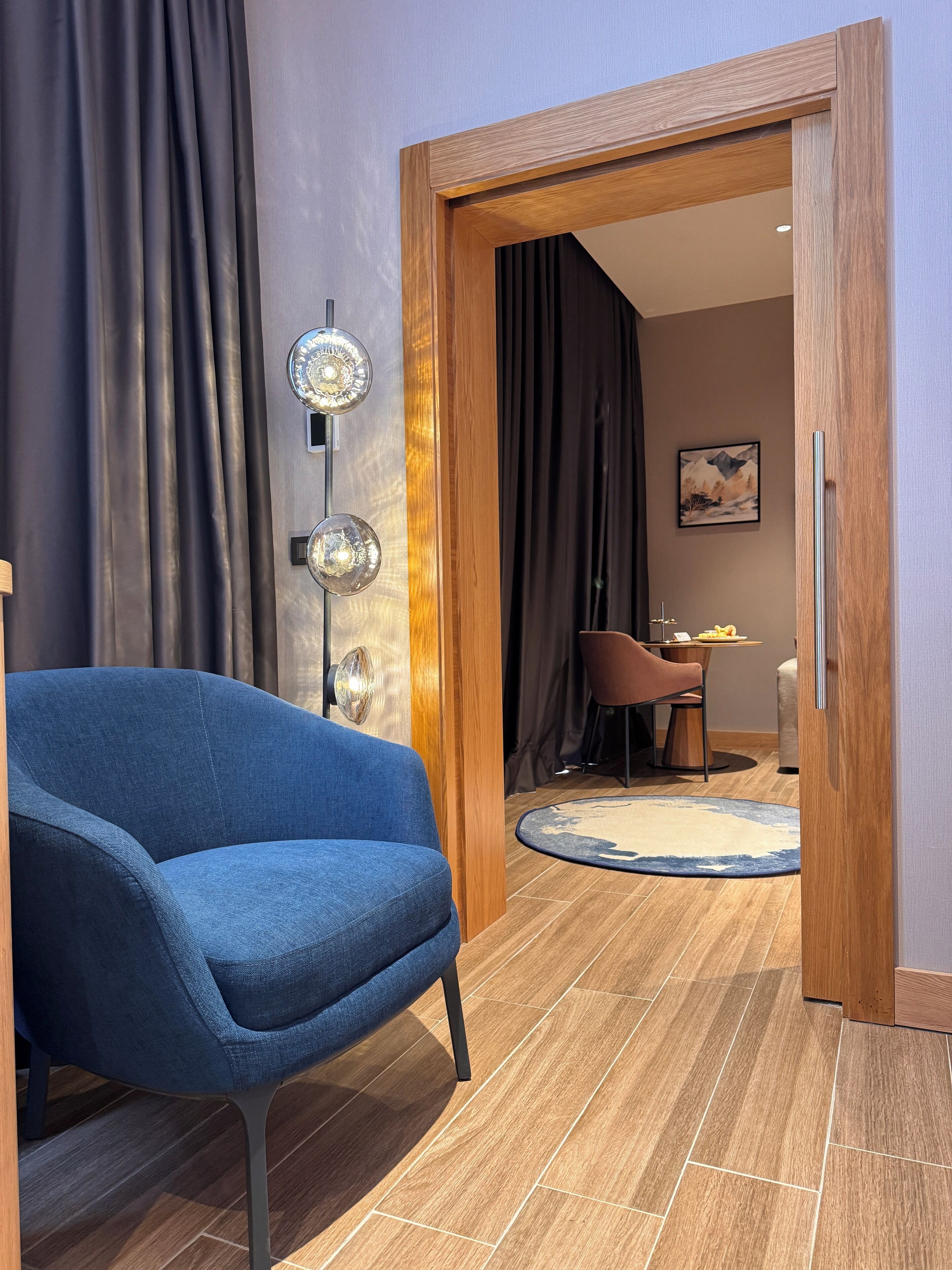 A modern room with a blue chair, wooden flooring, and a dining area visible through an open door.
