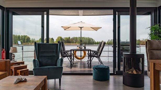 Un salon moderne doté d’une grande fenêtre donnant sur un lac, d’une table à manger avec des chaises, d’un canapé, d’une petite table avec une lampe et d’un poêle à bois.