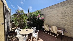 Um pátio com mesa redonda e cadeiras brancas, cercado por plantas e um muro de concreto.