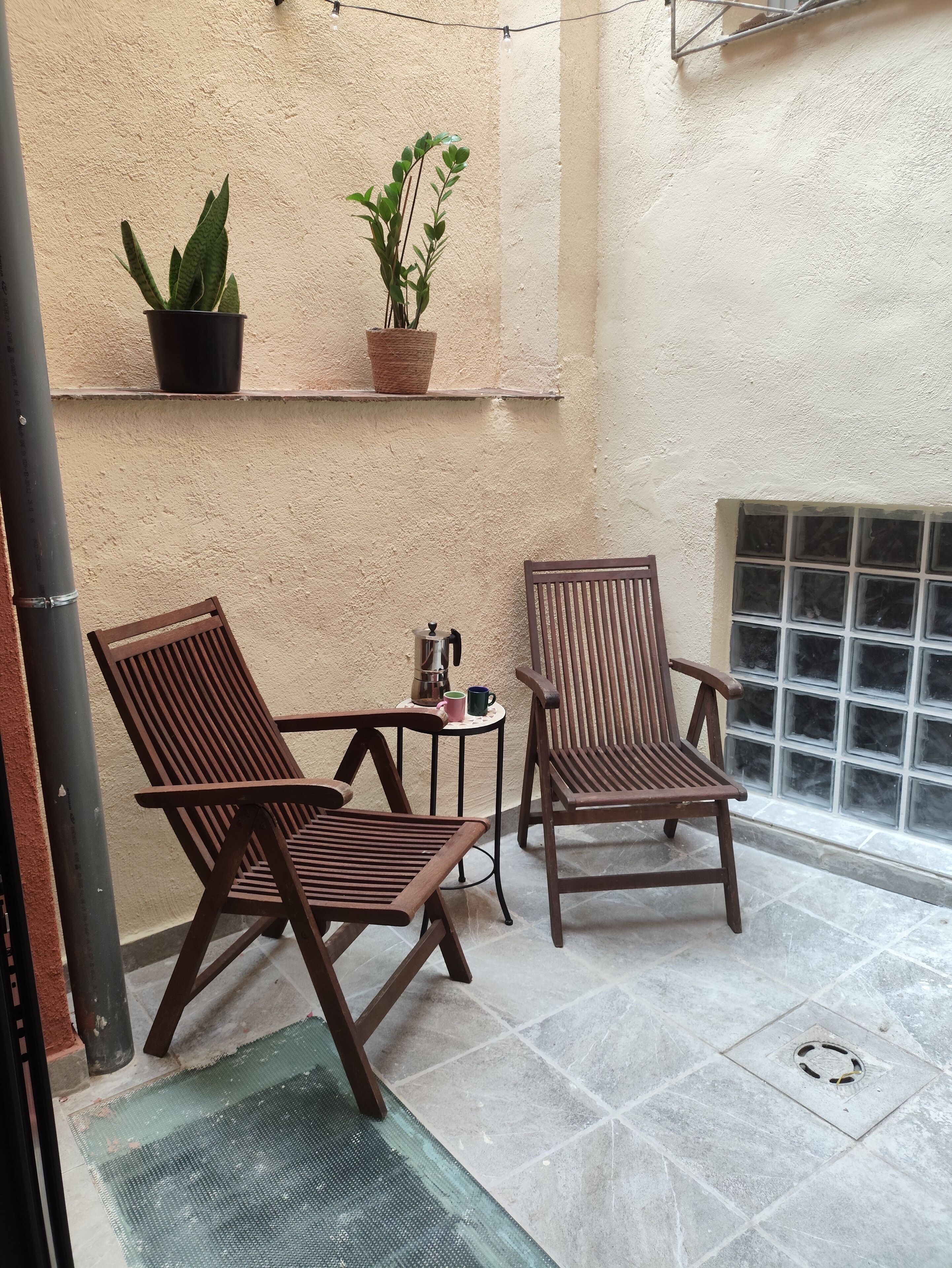 Una pequeña zona de estar al aire libre con dos sillas de madera y una mesa, una planta en maceta y una ventana con un diseño de cuadrícula.