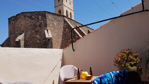 Un balcón con una mesa, sillas y una planta en maceta. Se divisa un edificio de piedra con una torre al fondo.