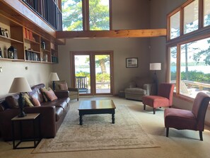 A spacious living room with a brown leather sofa, a wooden coffee table, and a red armchair. There is a bookshelf filled with books and decorative items, and a large window offering a view of the outdoors. - Lakefront 4-Bedroom Home on Lake Champlain with Stunning Views (Isle La Motte)