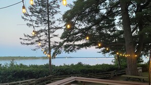Una terraza de madera con bancos que da a una masa de agua, luces colgantes y un árbol grande.
