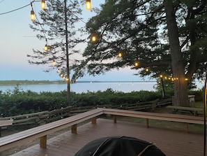 A wooden deck with benches overlooking a body of water, string lights, and a large tree. - Lakefront 4-Bedroom Home on Lake Champlain with Stunning Views (Isle La Motte)