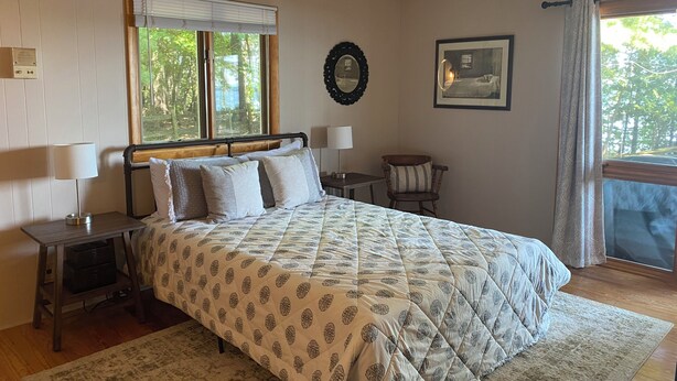 Un dormitorio con cama, mesitas de noche, una silla, ventana con persianas, un cuadro enmarcado y vistas al exterior.