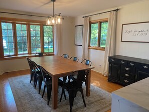 A dining area with a wooden table and chairs, large windows with curtains, a map on the wall, and a black cabinet with drawers. - Lakefront 4-Bedroom Home on Lake Champlain with Stunning Views (Isle La Motte)