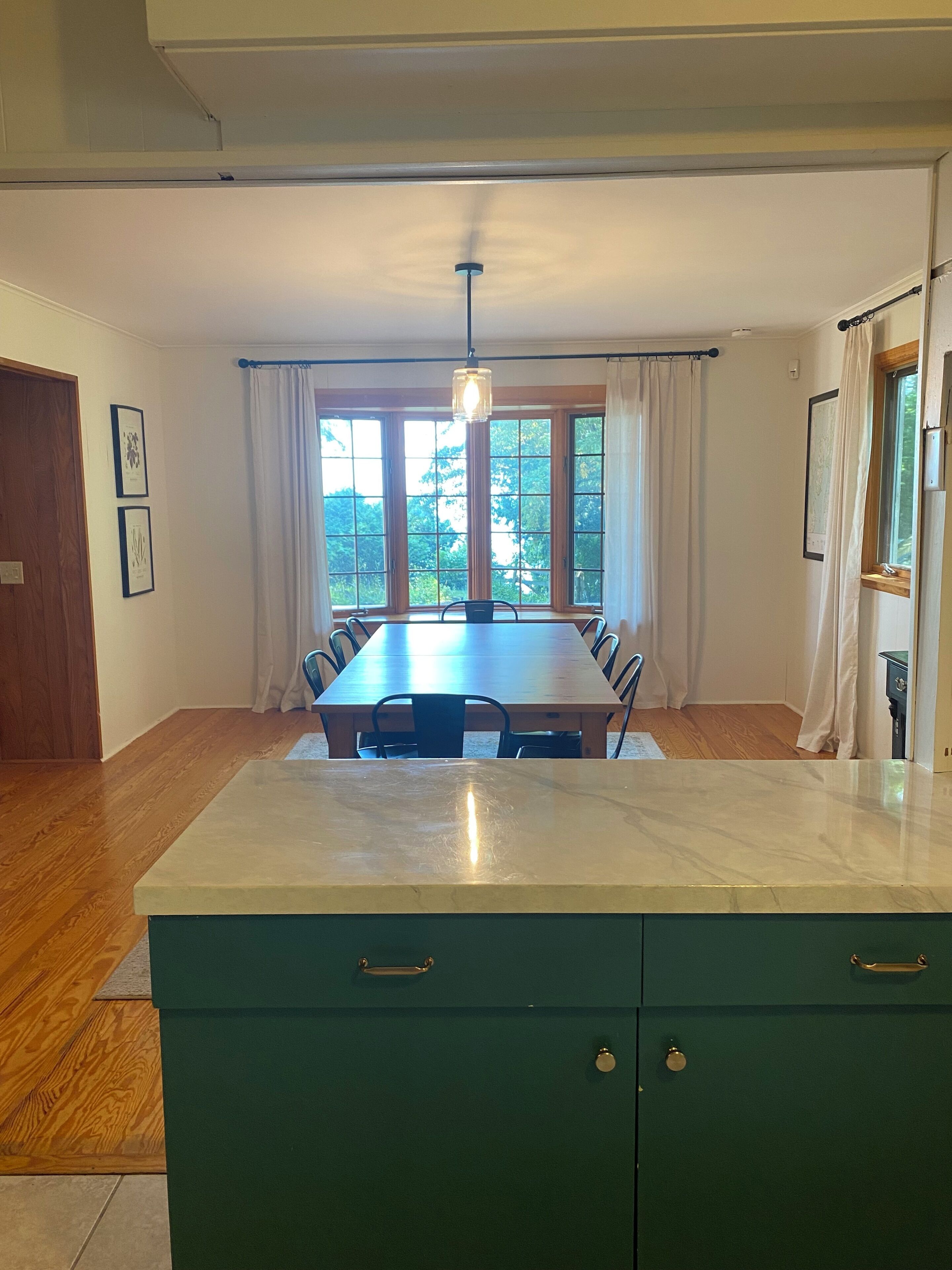 A dining area with a long table, chairs, a hanging light fixture, and a view through large windows.