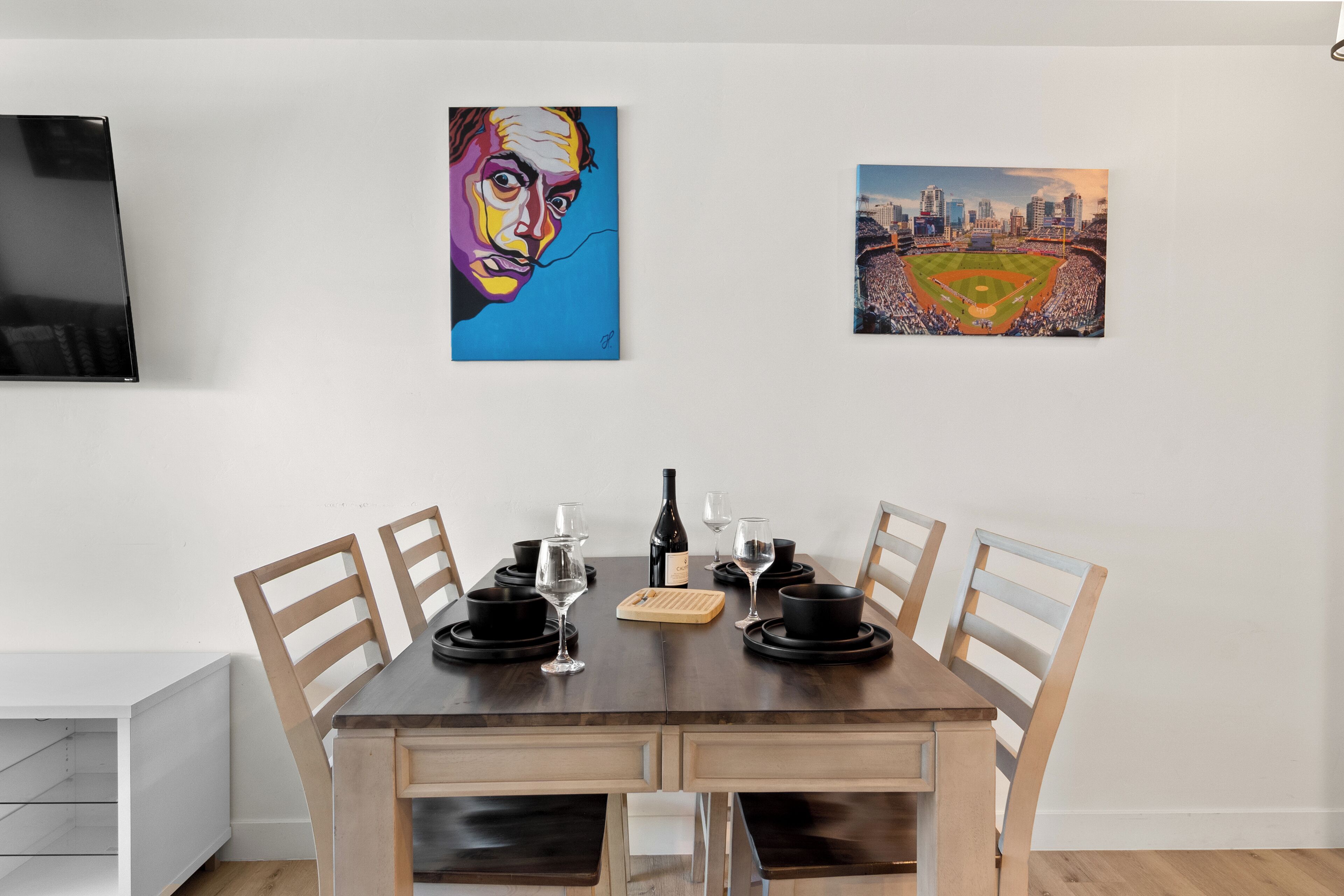 Un comedor con una mesa de madera dispuesta para cuatro personas, un televisor montado en la pared y dos cuadros sobre la mesa.