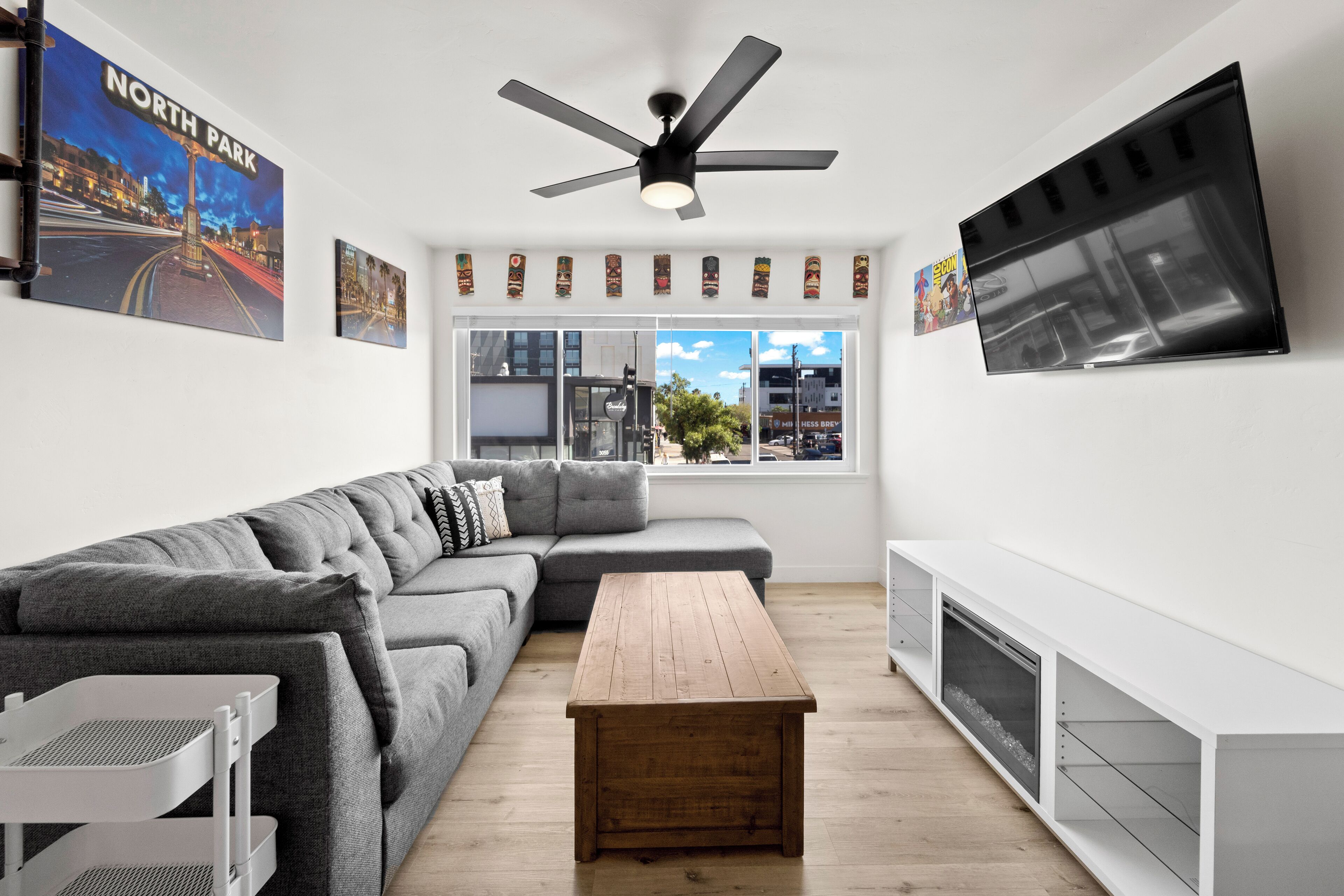 A modern living room with a grey sectional sofa, a wooden coffee table, a ceiling fan, and a TV mounted on the wall.