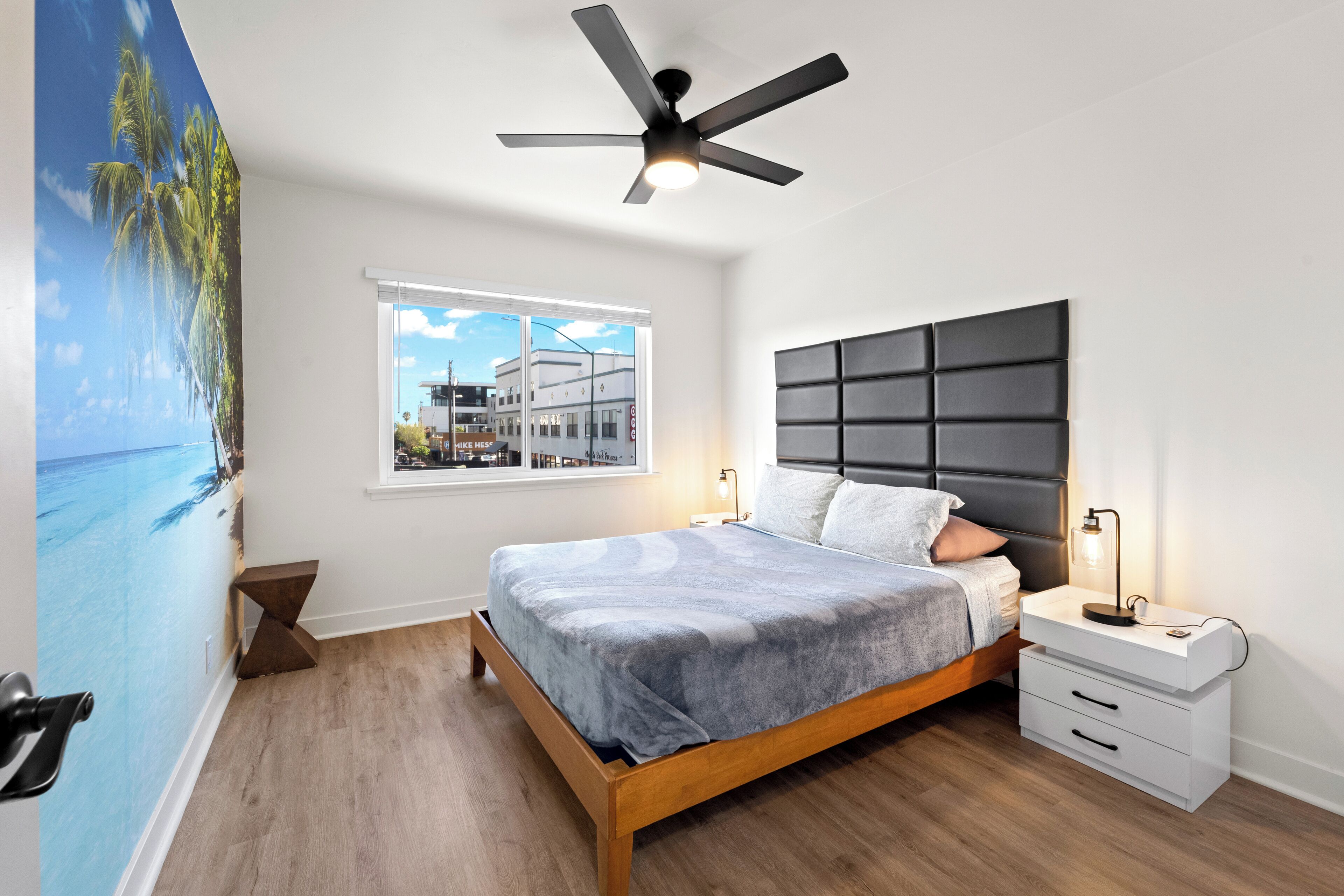 Un dormitorio con cama, mesita de noche, ventilador de techo, ventana con vistas a edificios y un mural en la pared.
