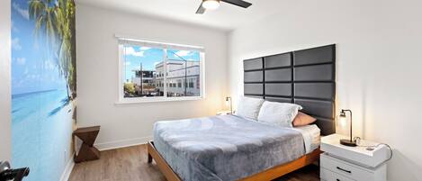 Un dormitorio con cama, mesita de noche, ventilador de techo, ventana con vistas a edificios y un mural en la pared.