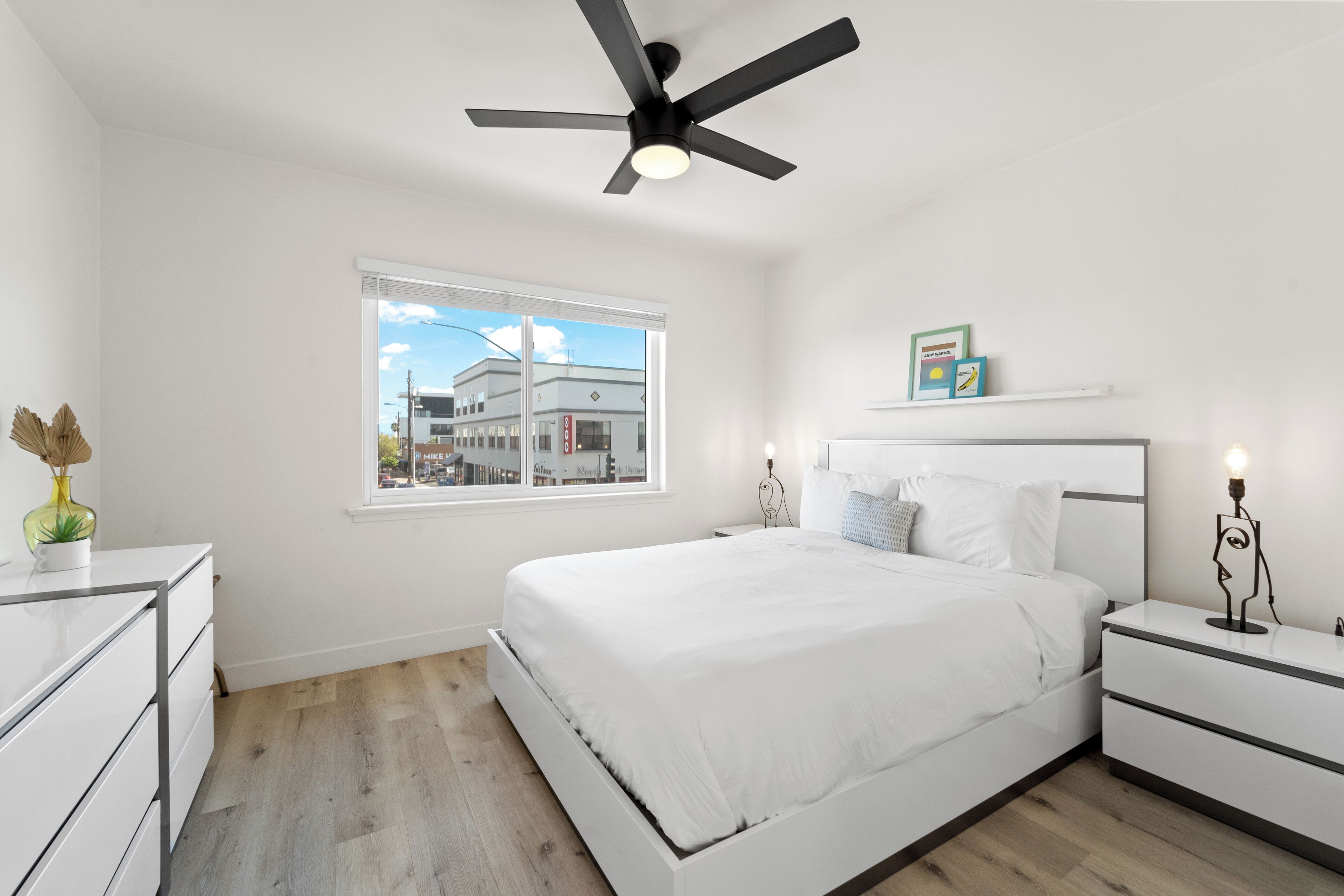 Un dormitorio moderno con una cama grande, un ventilador de techo, una mesita de noche con lámpara y una ventana con vista a la ciudad.