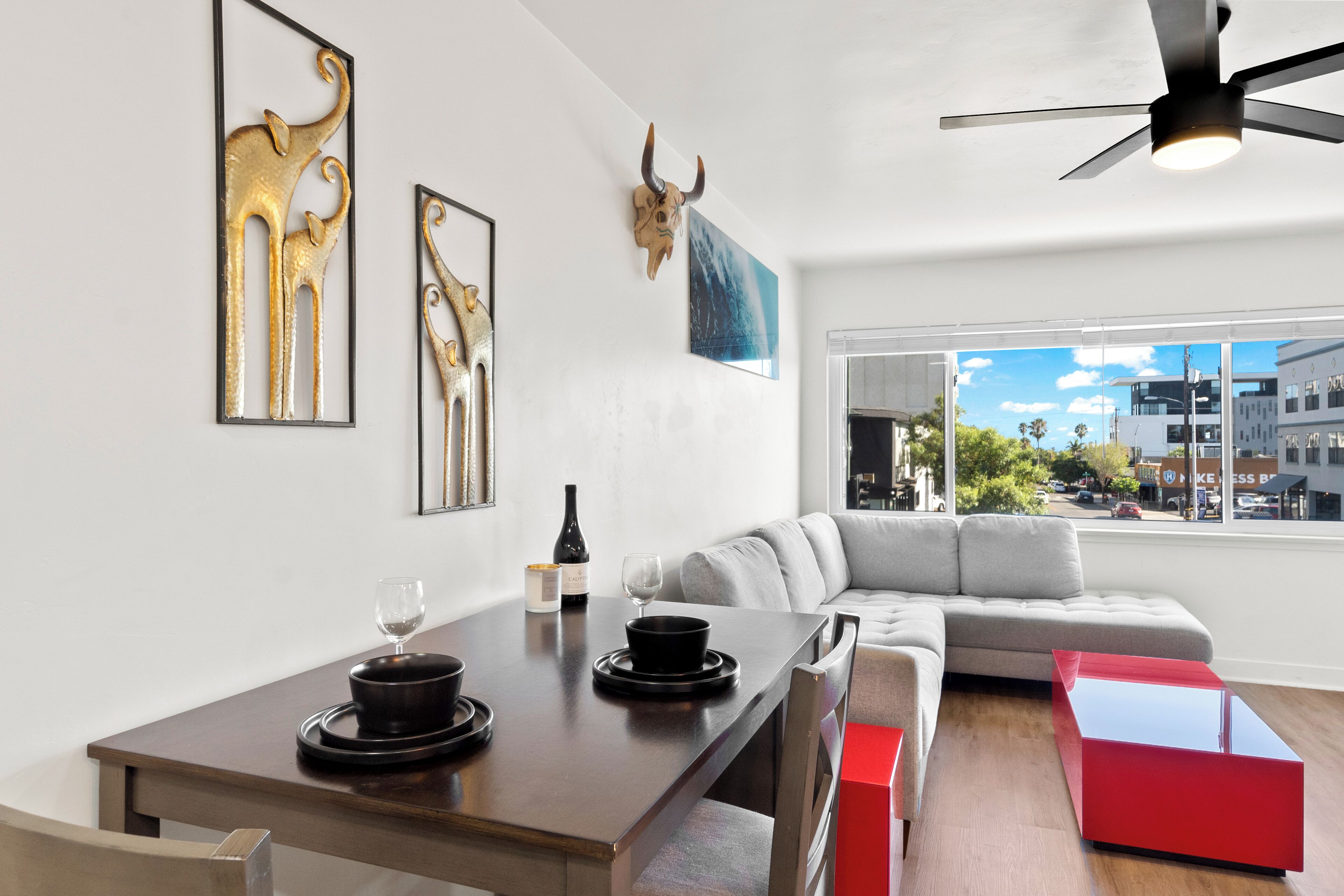 A modern living room with a dining area, a large window with a city view, a ceiling fan, and a bull skull decoration on the wall.