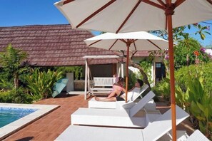 A poolside area with lounge chairs and umbrellas, a covered seating area, and a building with a tiled roof in the background. - Darya Sea View (Bali)