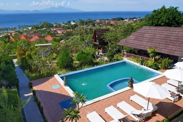 A swimming pool with a view of the ocean, surrounded by greenery and a building with a tiled roof.