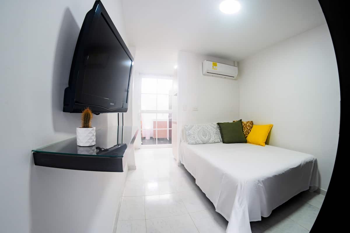 A hotel room with a bed, a TV mounted on a glass shelf, an air conditioning unit, and a view of the outside.