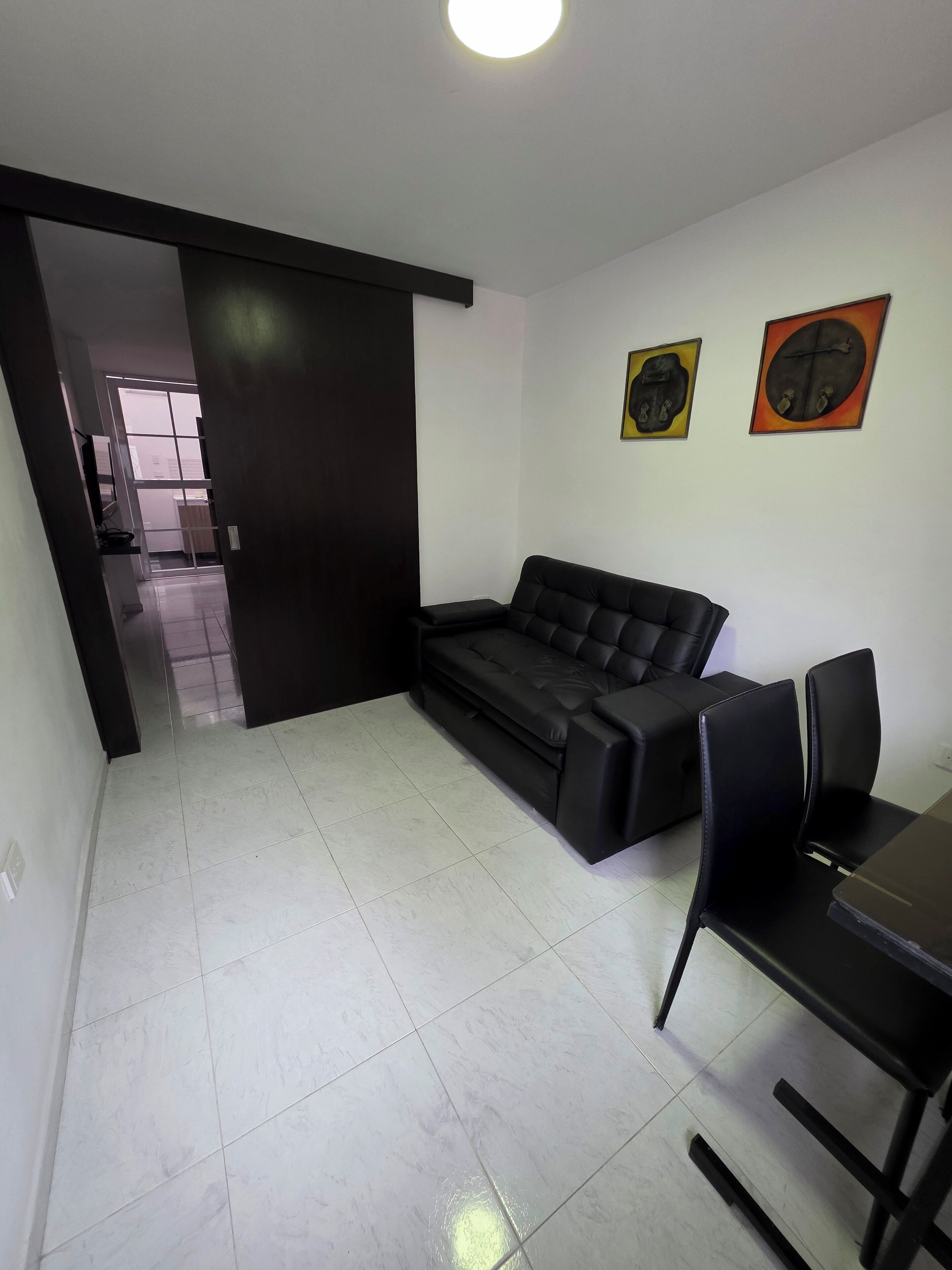 Una sala de estar moderna con un sofá negro, una mesa de comedor con sillas y dos cuadros abstractos en la pared.
