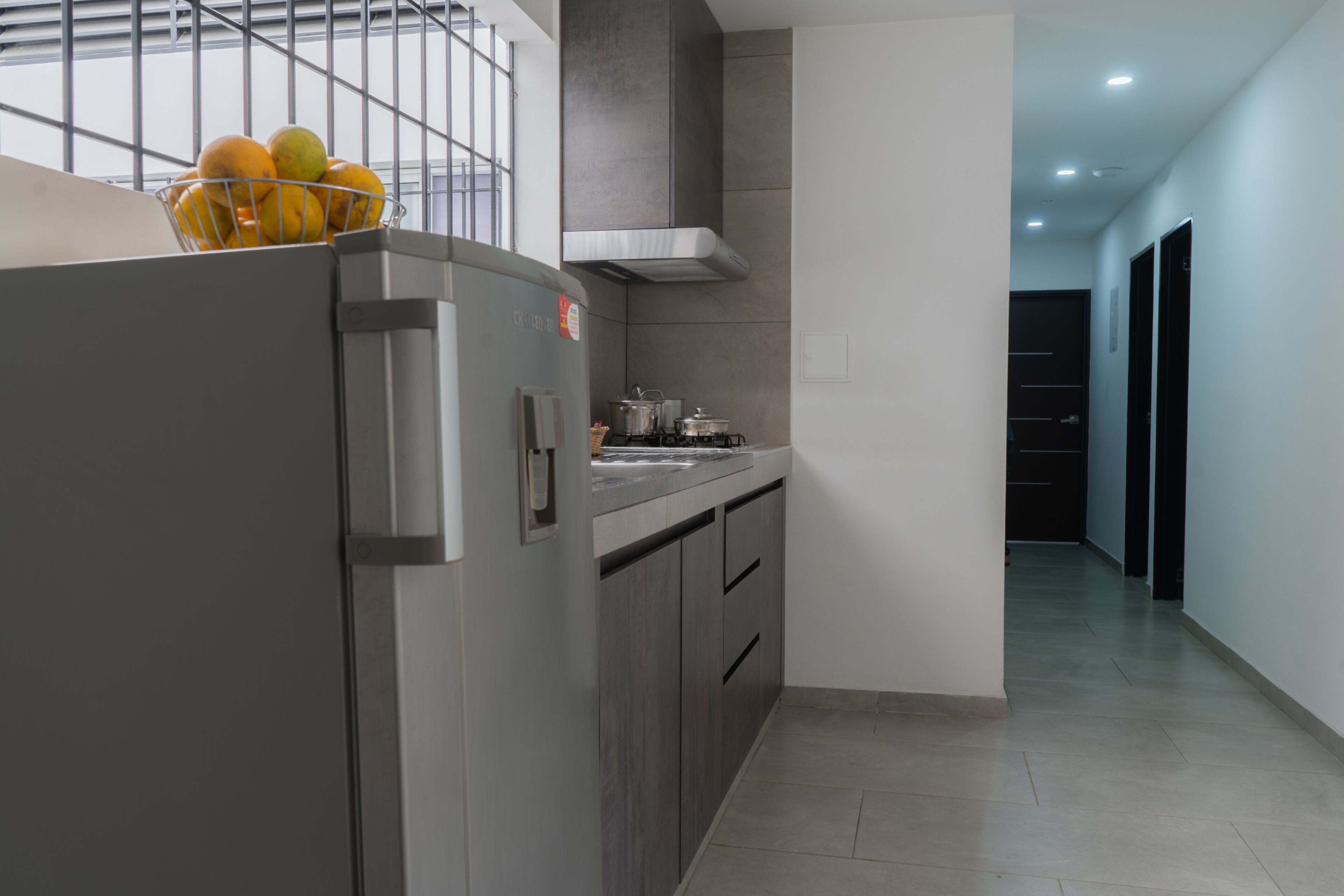 Una cocina moderna con un refrigerador de acero inoxidable, un lavamanos y una estufa. Hay una ventana con una reja de metal y una cesta de fruta en la encimera.